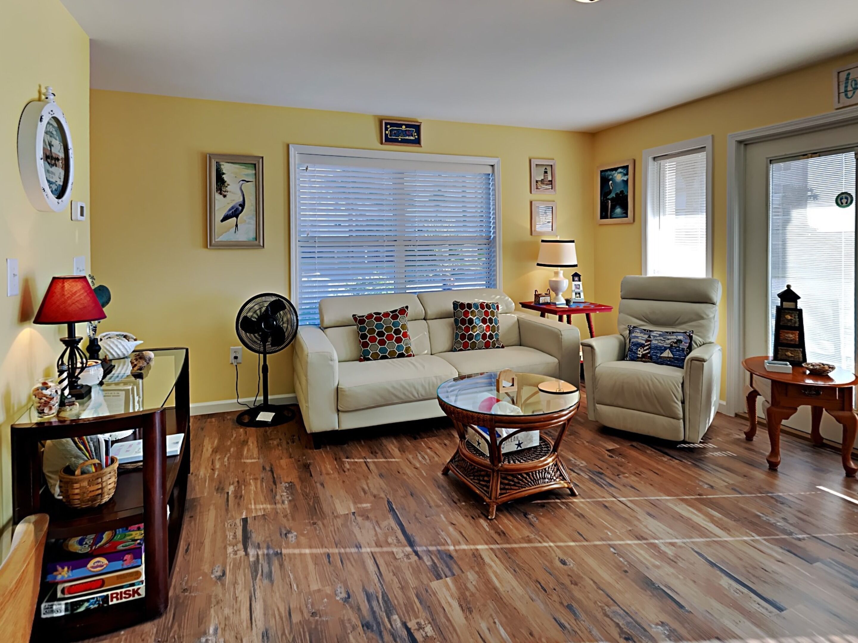 A living room with a beige sofa, a coffee table, and a fan. There are two windows with blinds, a lamp, and a clock on the wall.
