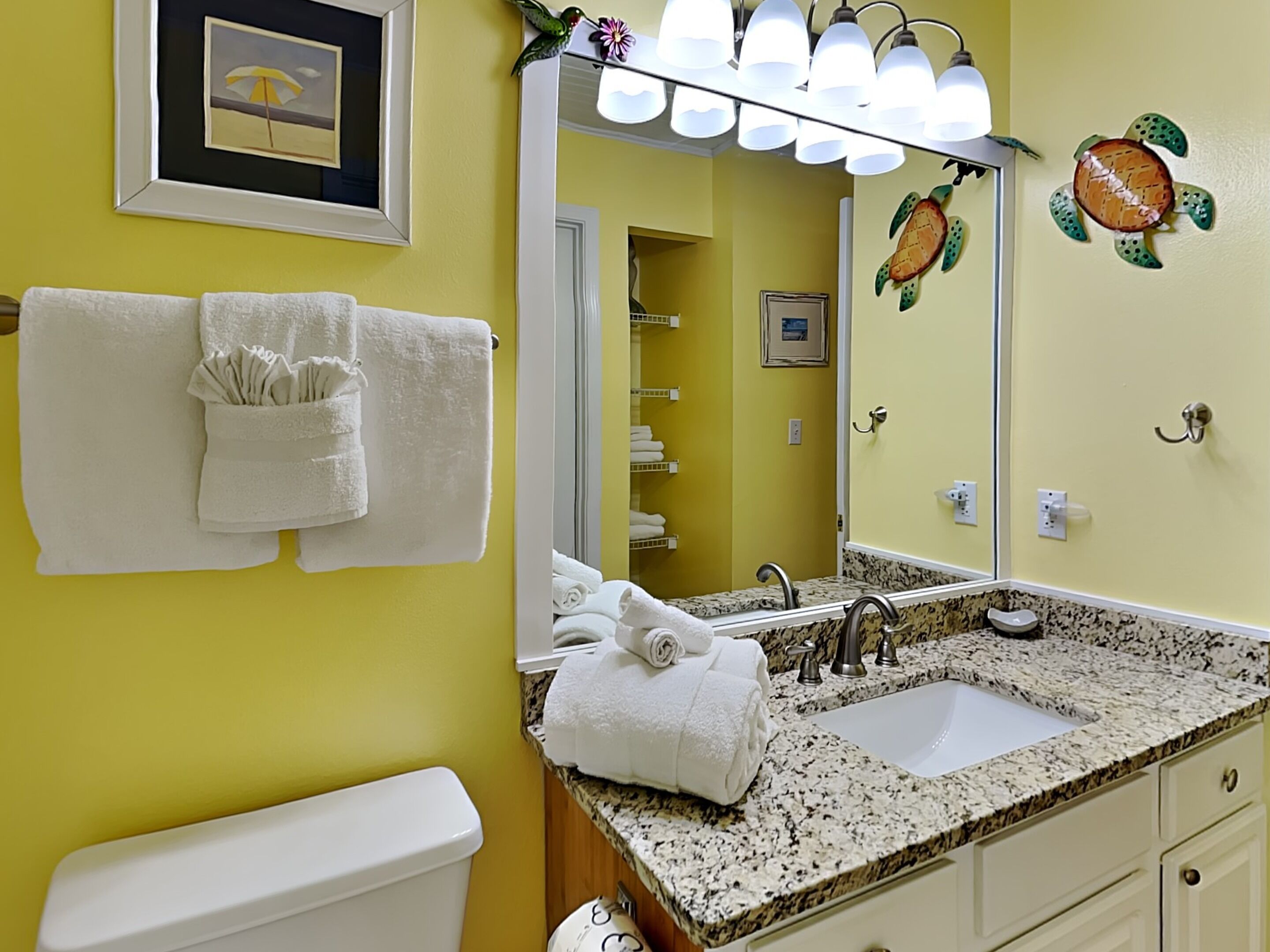 A bathroom with a yellow wall, a white sink, and a mirror. There are towels hanging on the wall and a framed picture above the mirror.