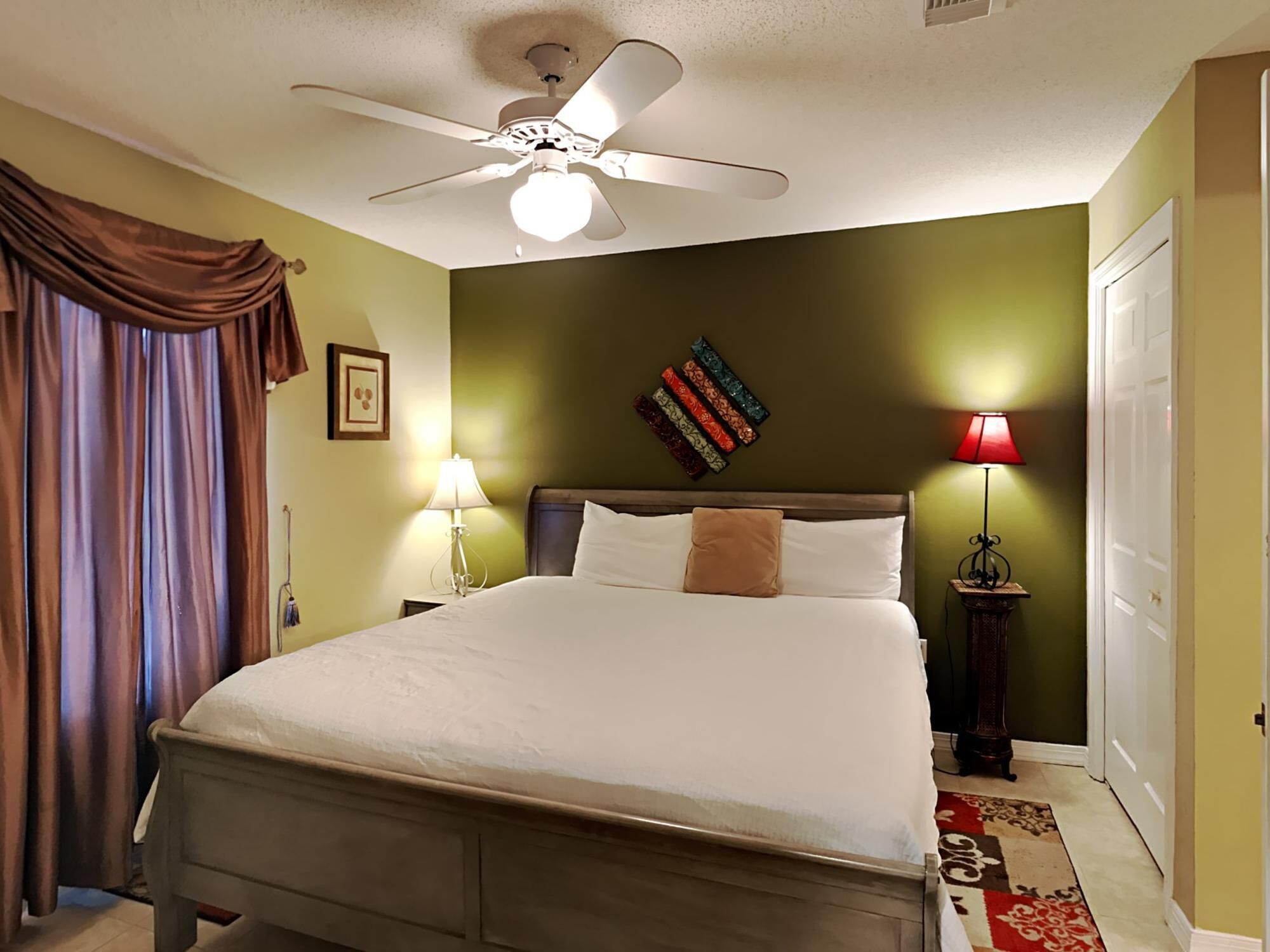Une chambre à coucher avec un lit, un ventilateur de plafond, deux lampes de chevet, une horloge murale et une porte.