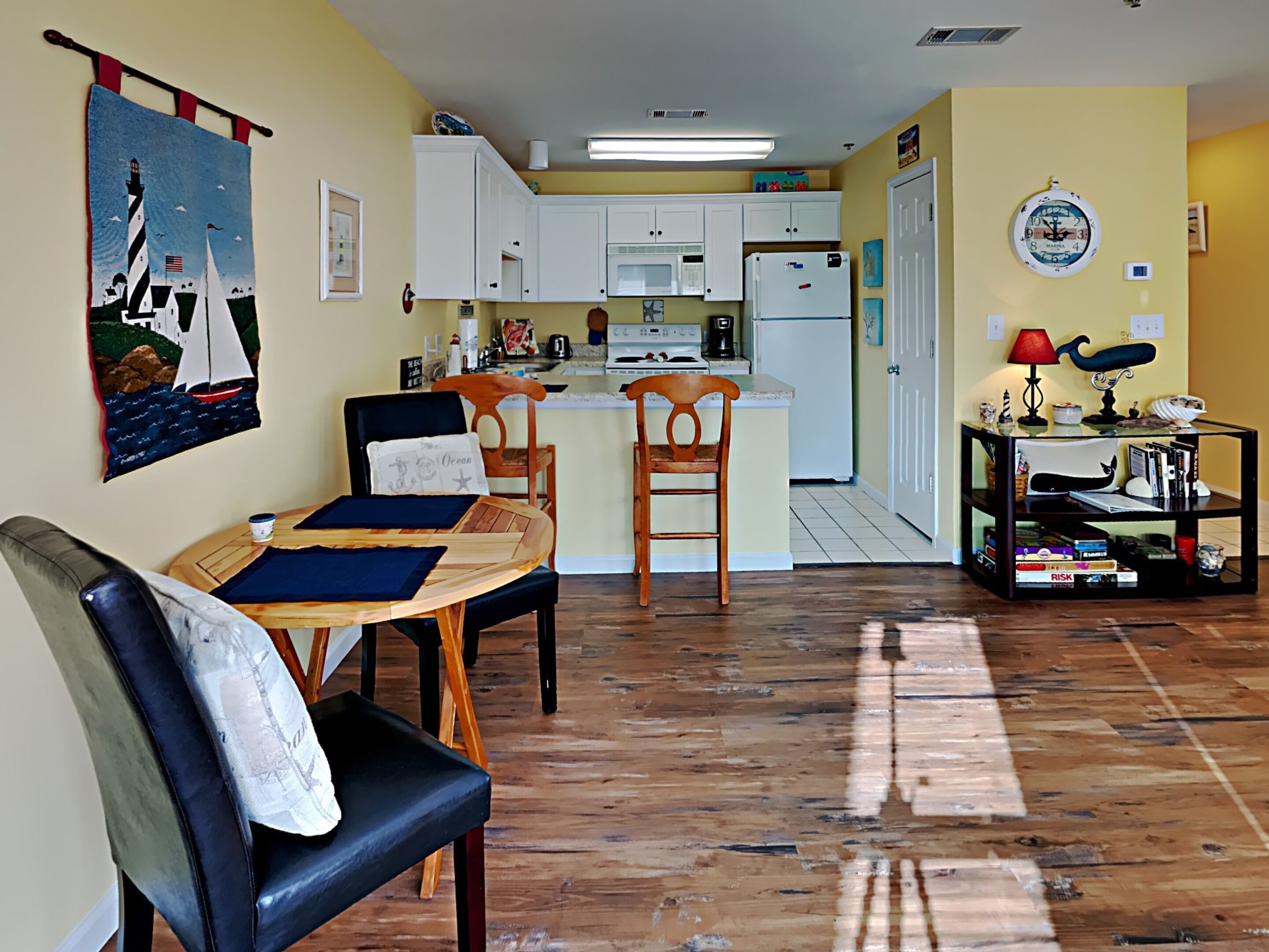 A compact kitchen with white cabinets, a refrigerator, and a microwave. There is a dining area with a wooden table and chairs, and a small bookshelf with books and decorative items.