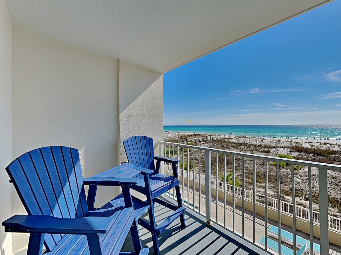 Een balkon met uitzicht op het strand, een zwembad en blauwe Adirondack-stoelen.