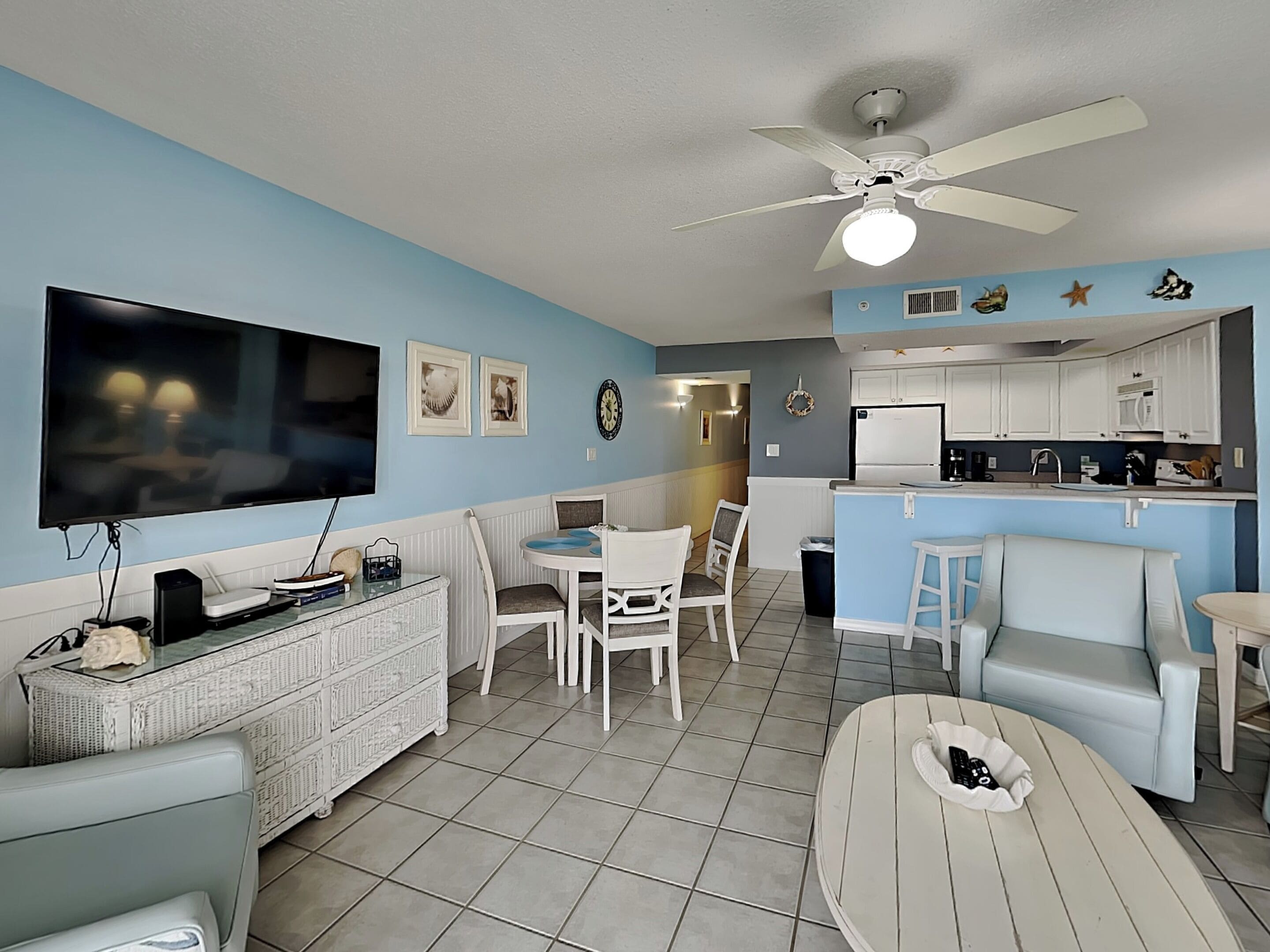 A living room with a TV, white furniture, a ceiling fan, and a kitchen area with white cabinets.