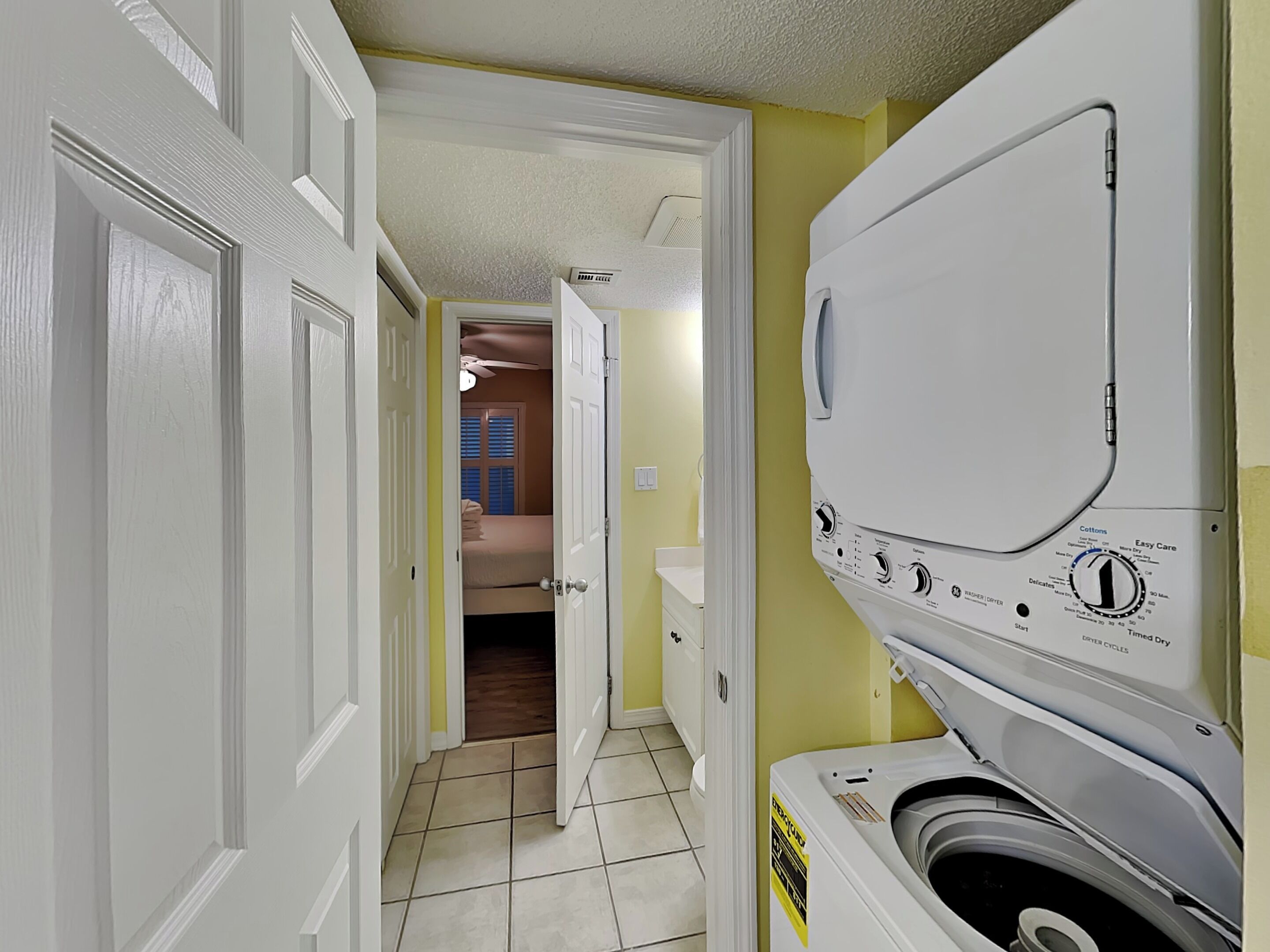A laundry room with a front-loading washing machine, a door leading to a bedroom, and a ceiling fan.