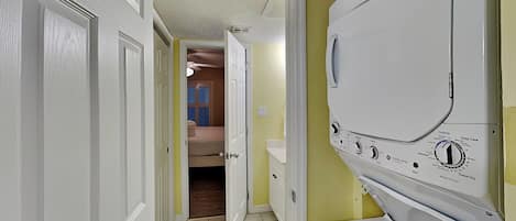 A laundry room with a front-loading washing machine, a door leading to a bedroom, and a ceiling fan.