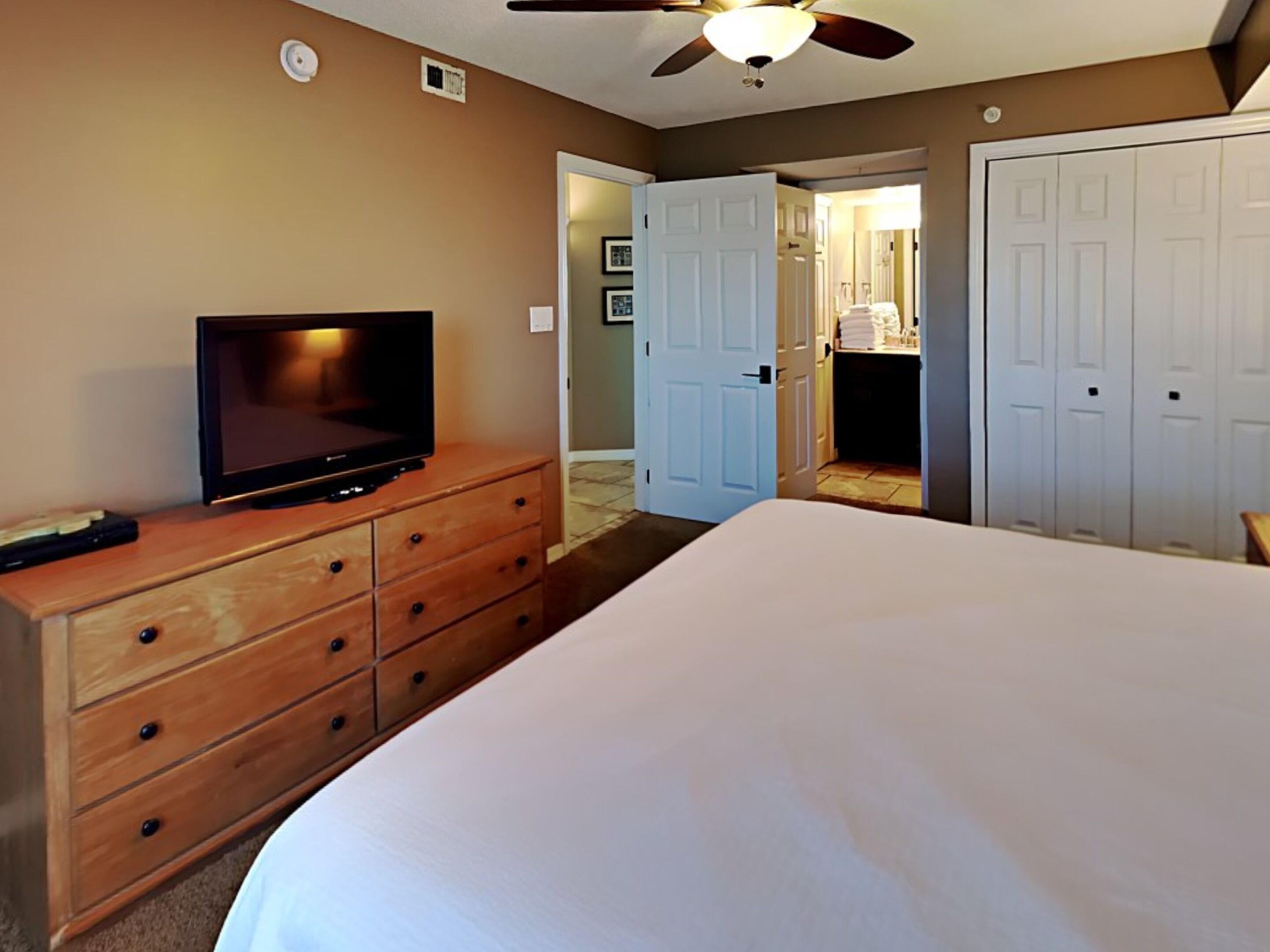 Habitación de hotel con cama, televisor sobre una cómoda de madera, ventilador de techo y baño visible a través de una puerta abierta.