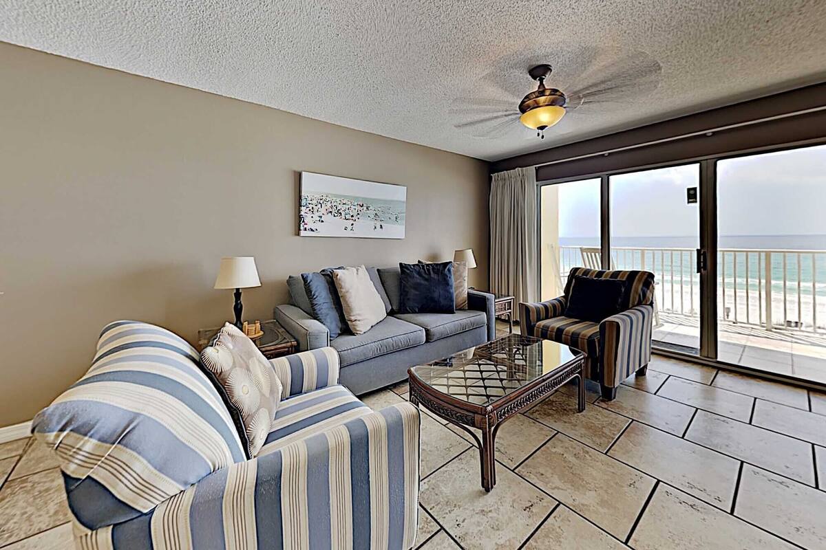 A living room with a sofa, armchair, and a glass coffee table. There is a ceiling fan, a painting on the wall, and a view of the ocean through large windows.