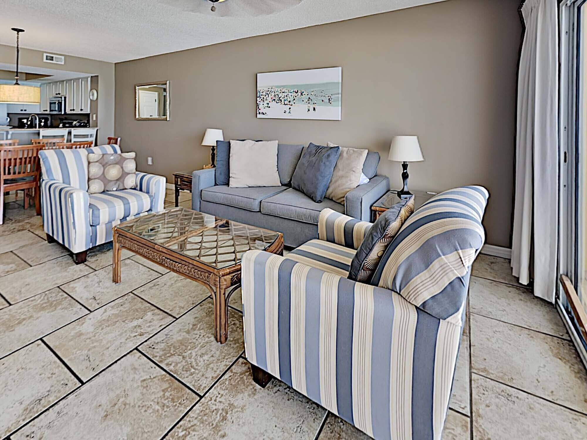 A living room with striped sofas, a glass coffee table, and a kitchen in the background.