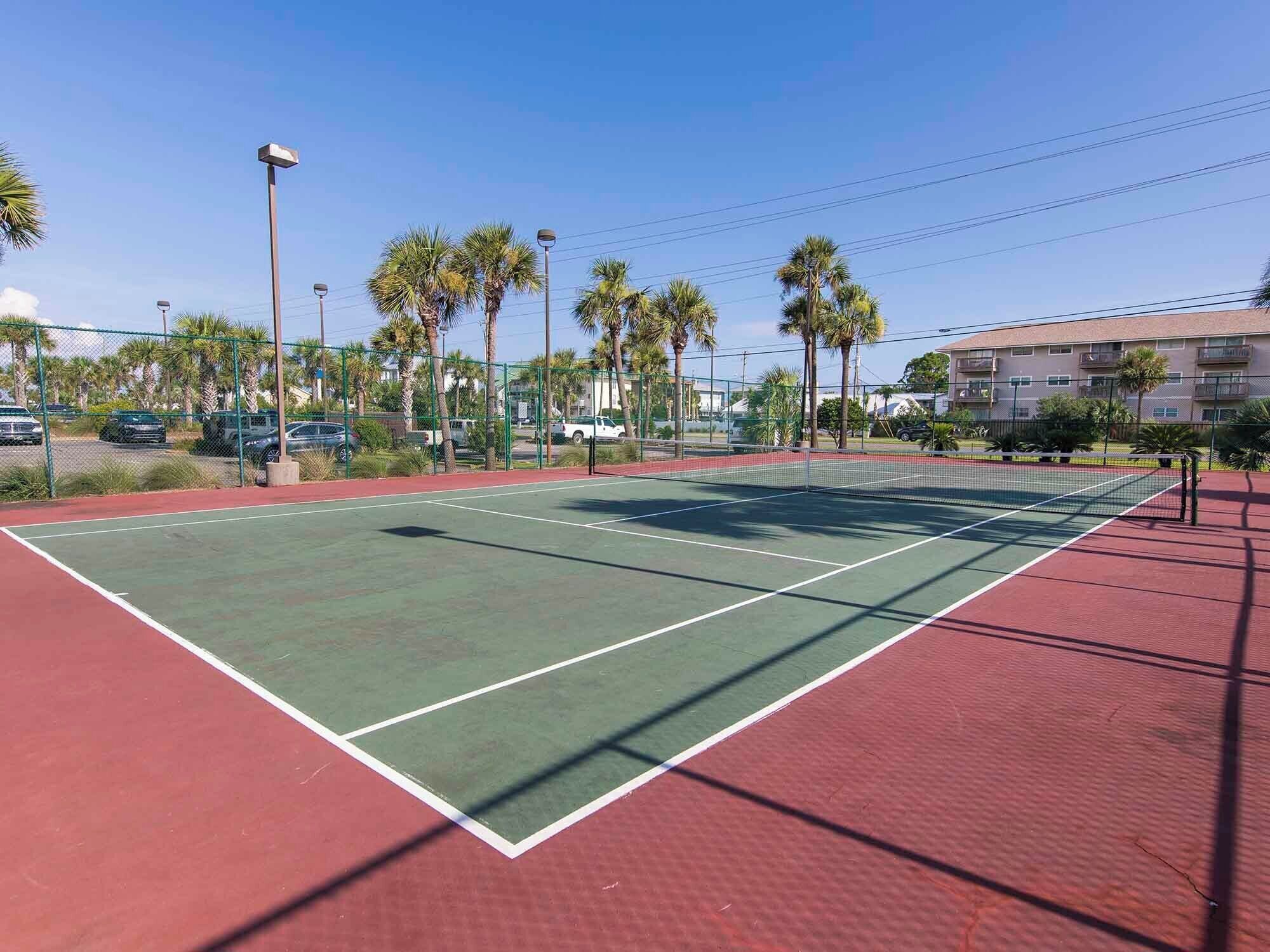 Ein Tennisplatz im Freien mit roter und grüner Belag, umgeben von einem Zaun und Palmen.