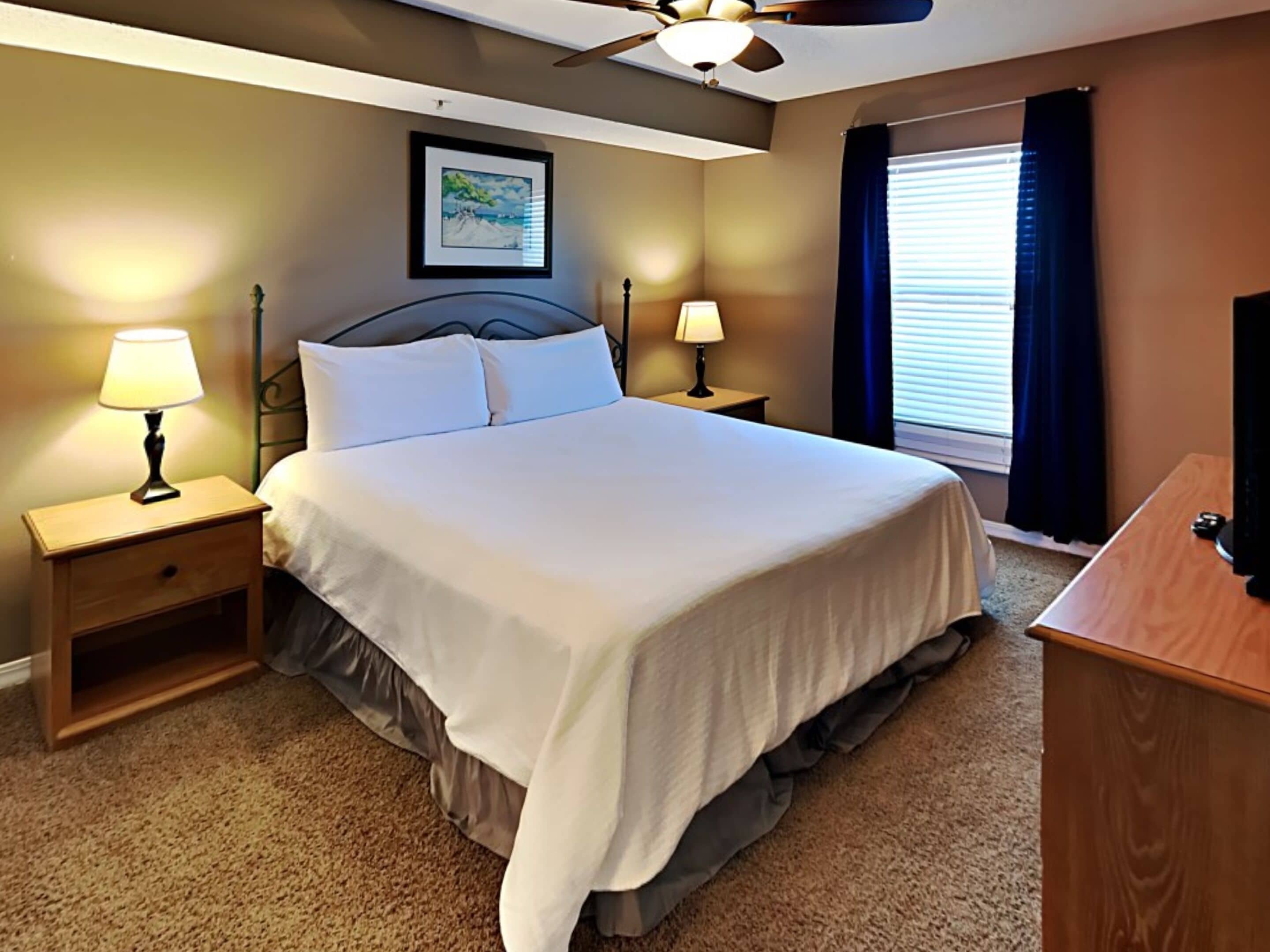 A neatly made bed in a hotel room with a ceiling fan, two bedside lamps, a framed picture on the wall, and a window with curtains.