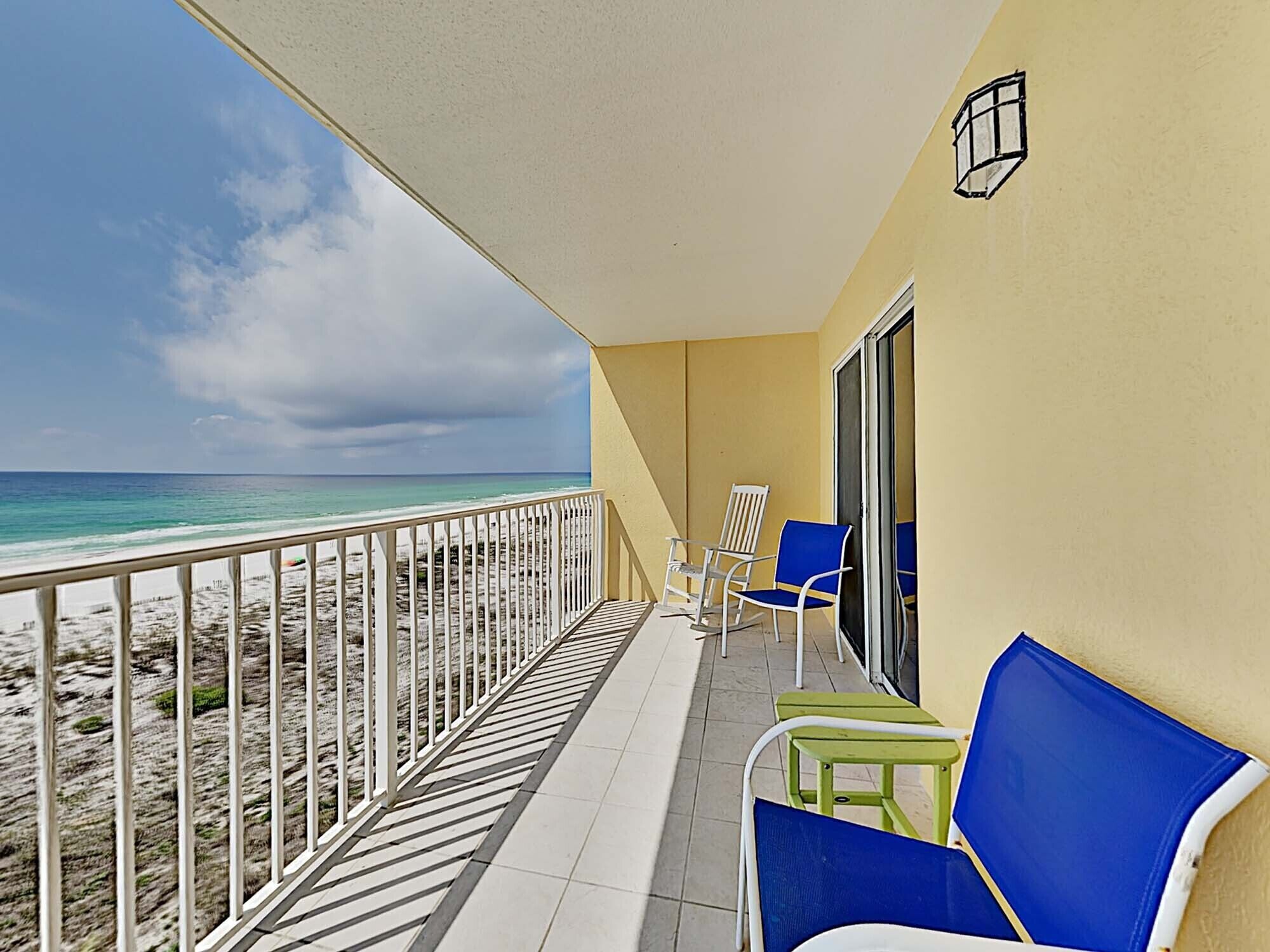 A balcony with a view of the beach, white railings, and blue and green chairs.