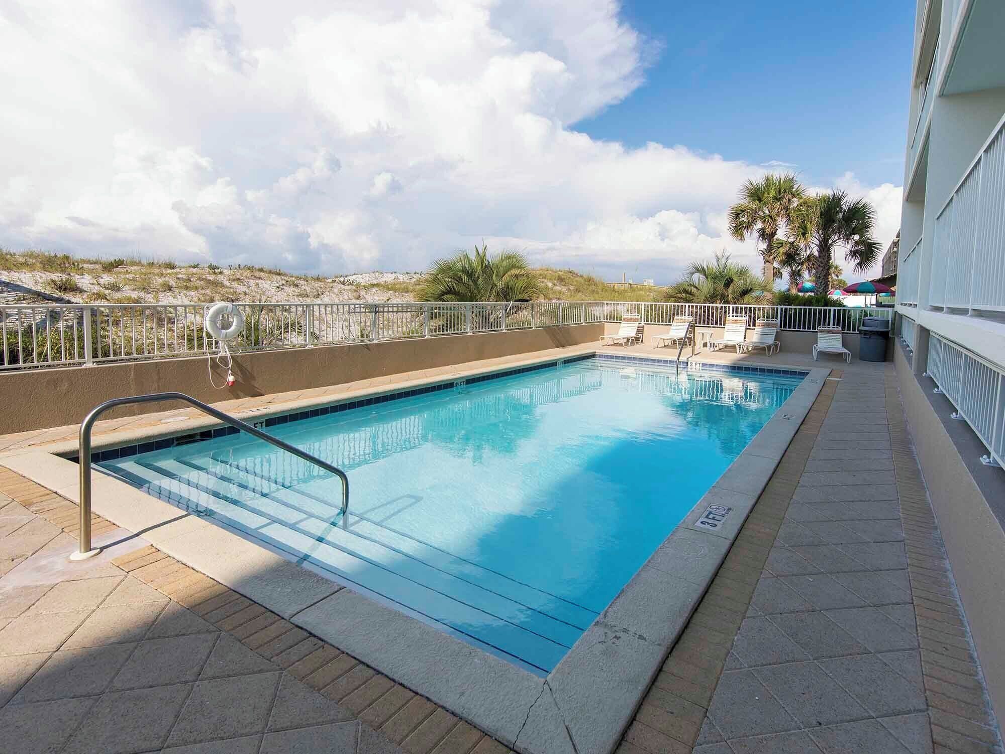 A rectangular swimming pool with a metal ladder, surrounded by a white fence. There are lounge chairs and umbrellas on the right side.