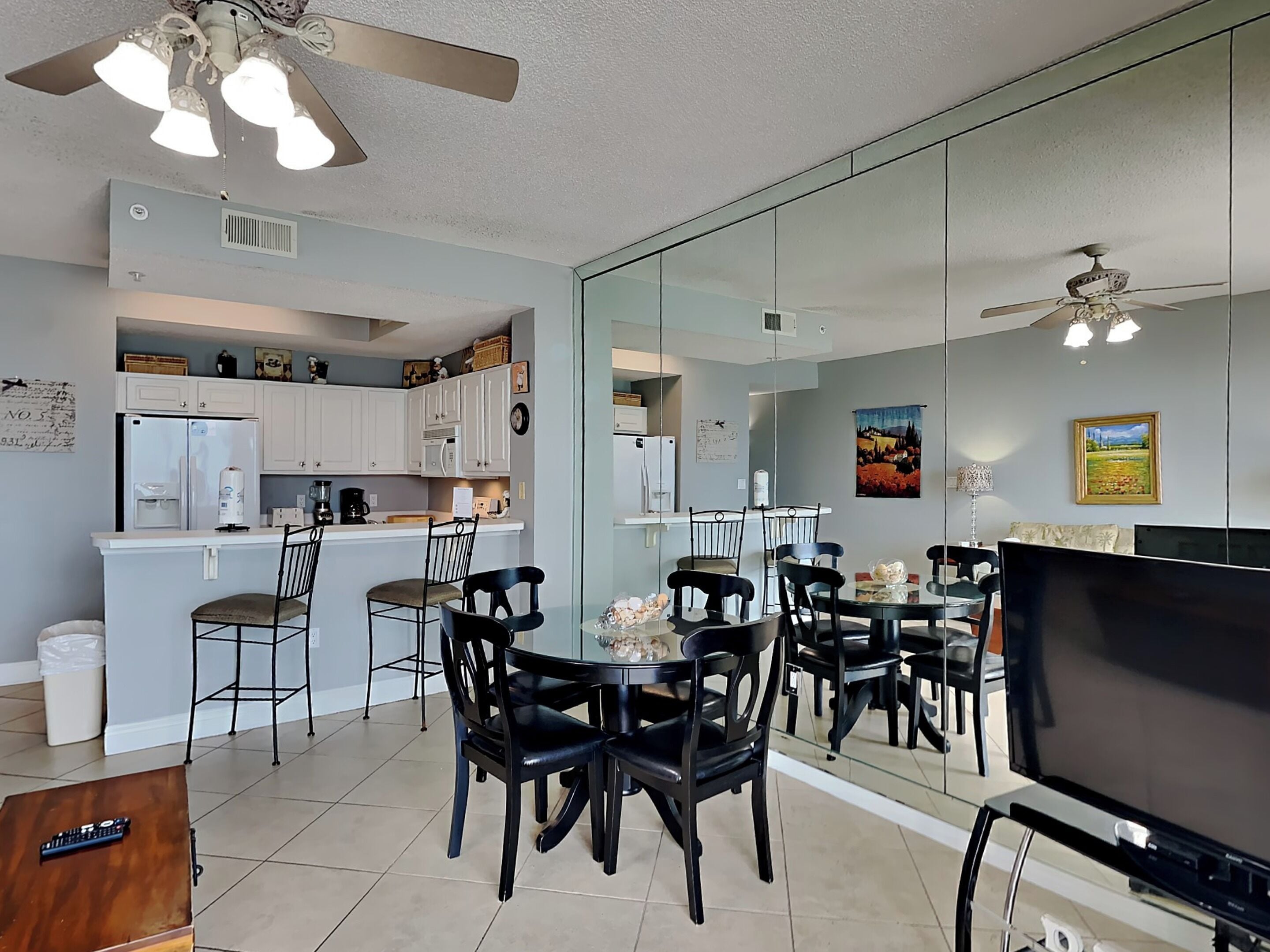 A modern kitchen with white cabinets, a refrigerator, and a coffee maker. There is a dining area with black chairs and a glass table. A TV is mounted on the wall.