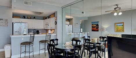 A modern kitchen with white cabinets, a refrigerator, and a coffee maker. There is a dining area with black chairs and a glass table. A TV is mounted on the wall.