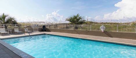 A rectangular swimming pool with clear blue water, surrounded by a tiled deck. There are lounge chairs and a small table on the deck.