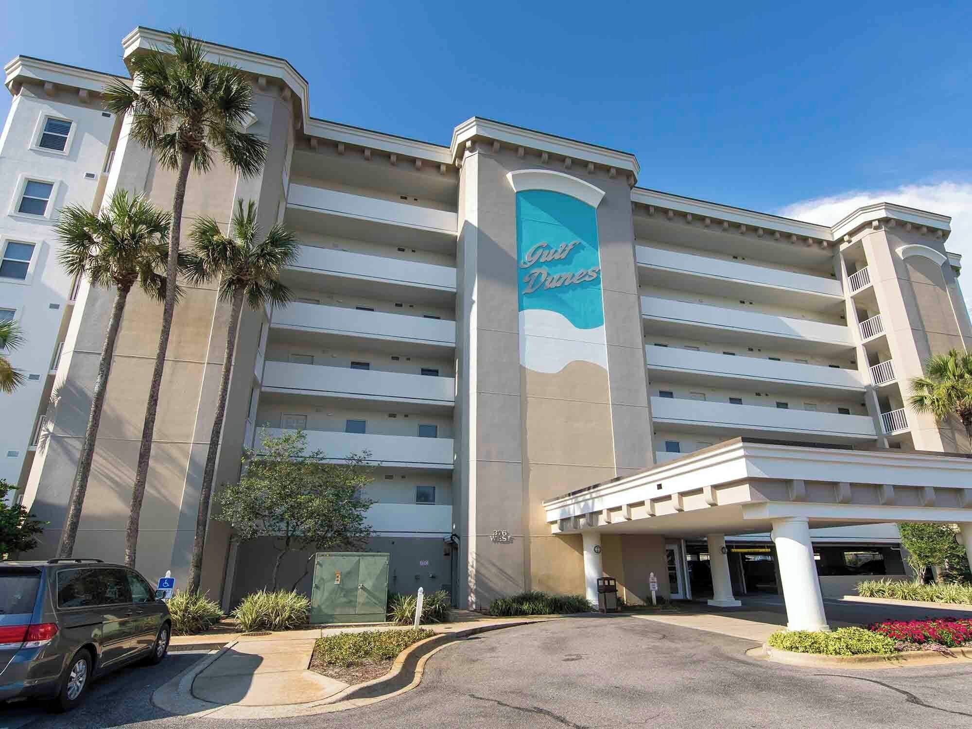 Un hotel de diverses plantes amb un cartell prominent que diu 'Gulf Dunes', amb un disseny arquitectònic modern, teulada plana i grans finestres.