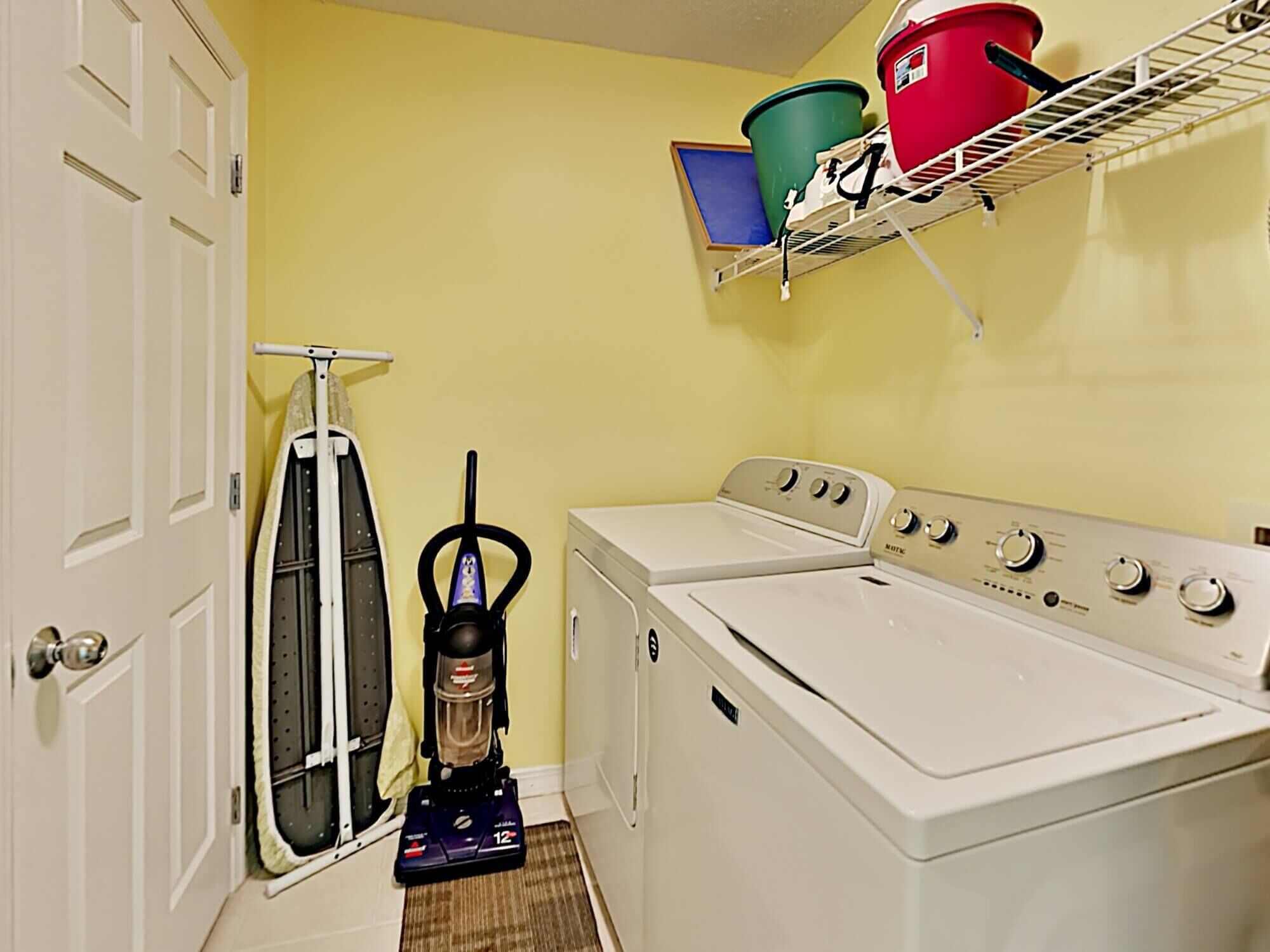 A laundry room with a front-loading washer and dryer, a vacuum cleaner, and a shelf with laundry baskets.