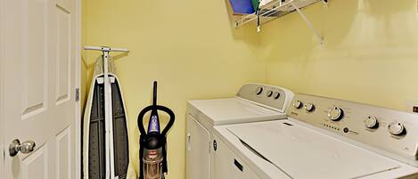 A laundry room with a front-loading washer and dryer, a vacuum cleaner, and a shelf with laundry baskets.