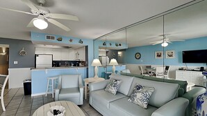 Ein modernes Wohnzimmer mit einer Couch, Sesseln, einem Couchtisch, einem Fernseher und einer Küche im Hintergrund.