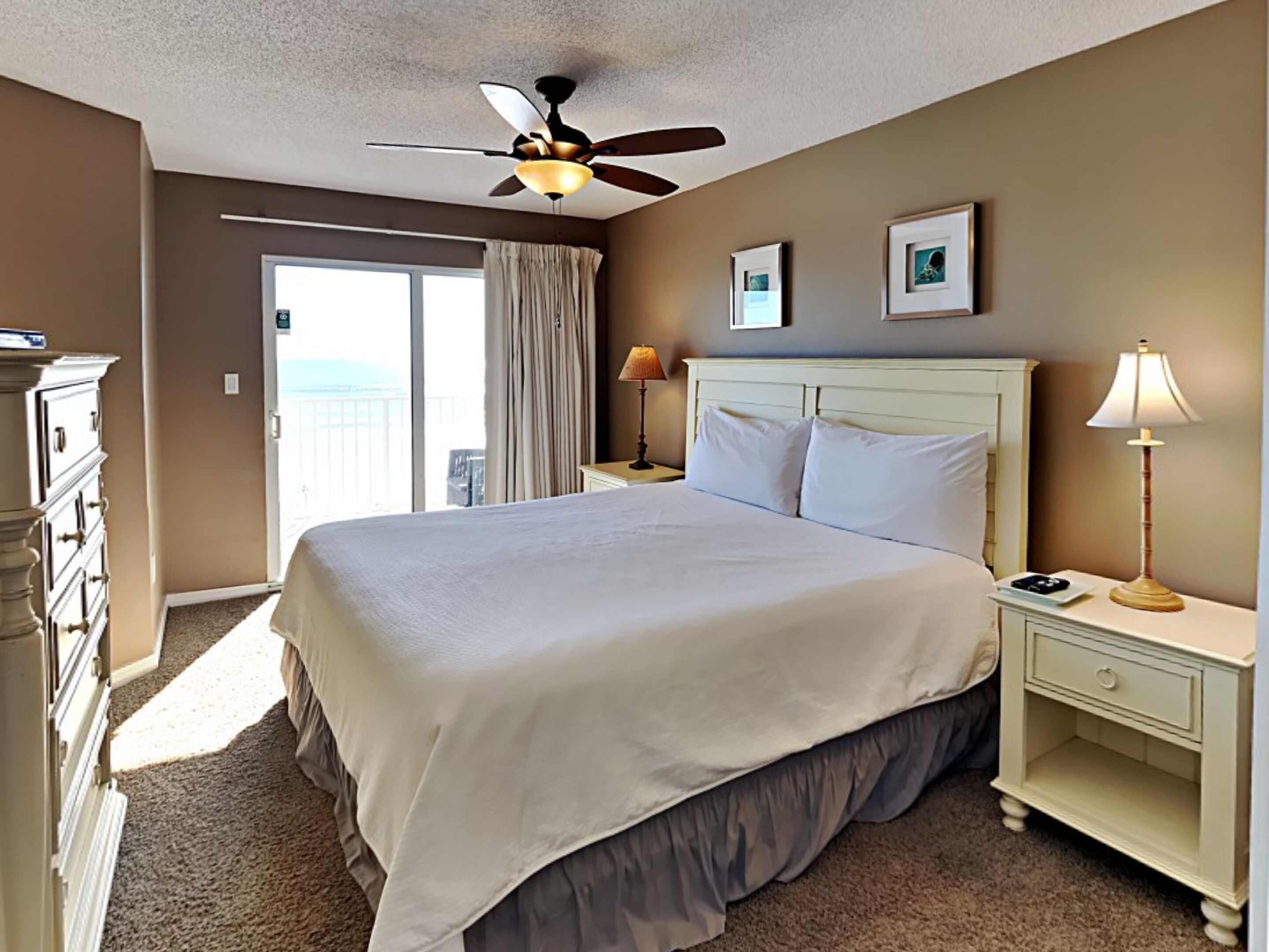 A hotel room with a bed, a ceiling fan, a nightstand with a lamp, a dresser, and a view of the outside.