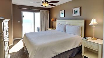 A hotel room with a bed, a ceiling fan, a nightstand with a lamp, a dresser, and a view of the outside.