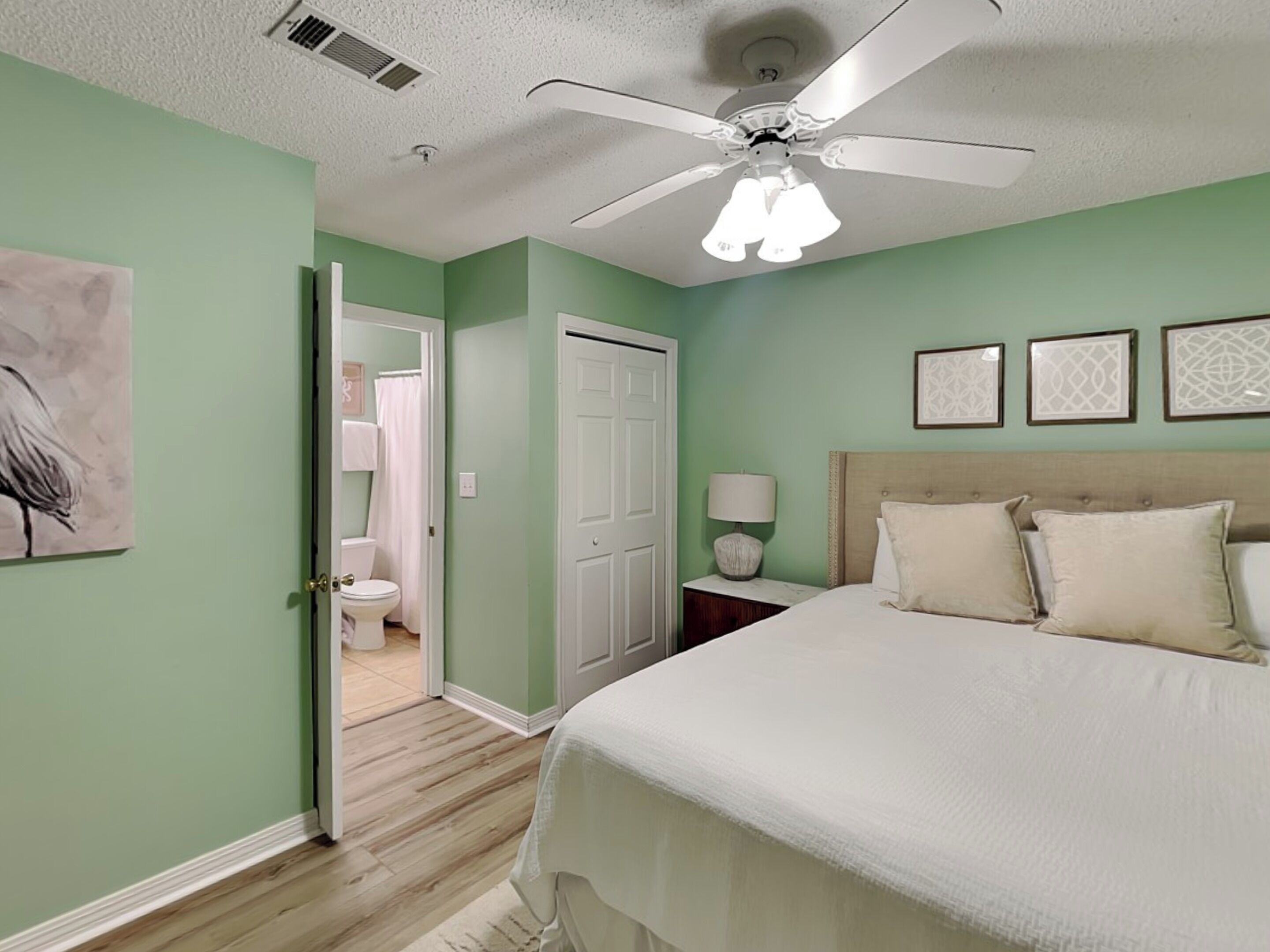 A bedroom with a bed, a nightstand, a ceiling fan, and a door leading to a bathroom.