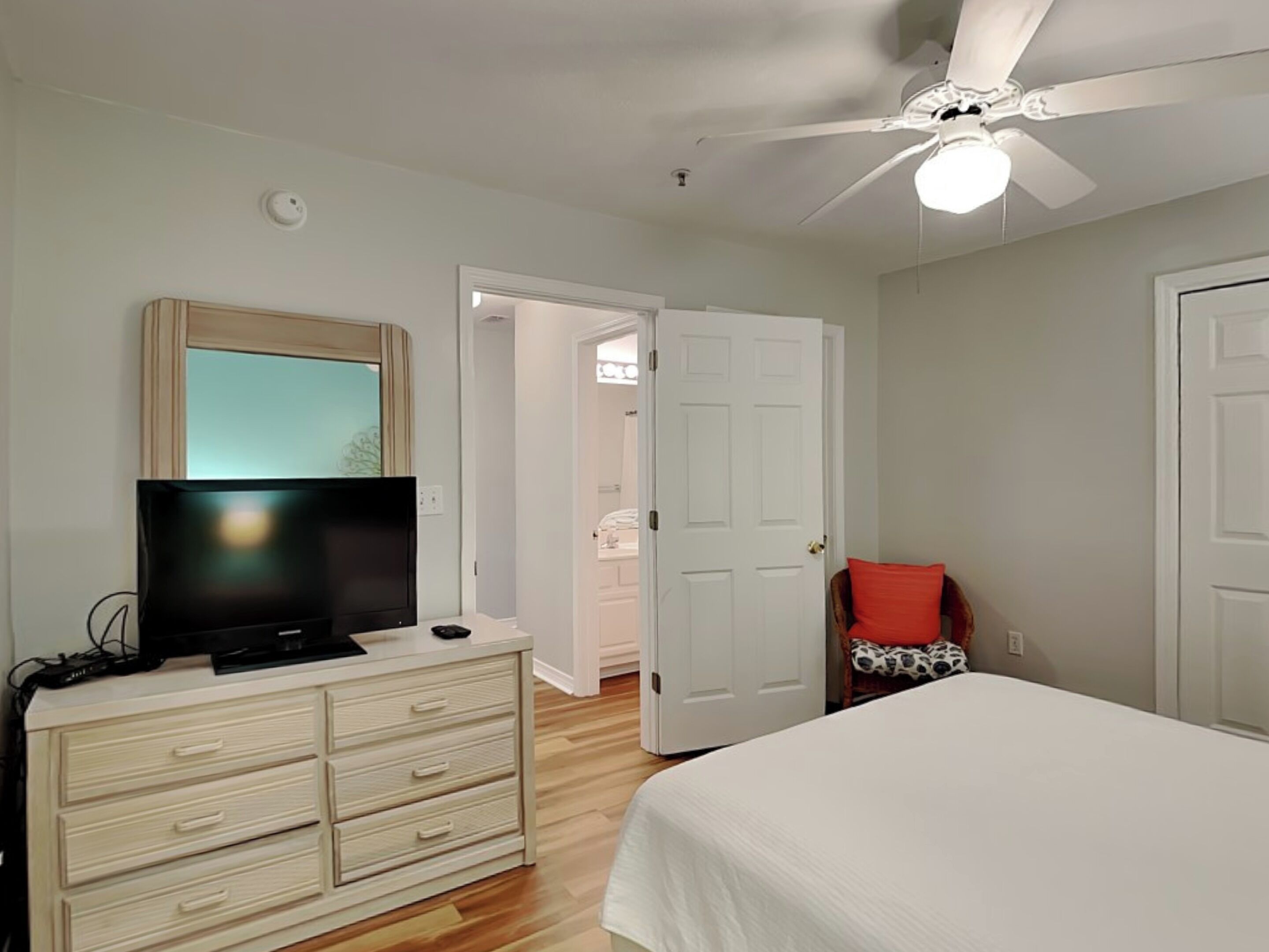 Quarto com cama, cômoda, televisão, ventilador de teto e porta para um banheiro.