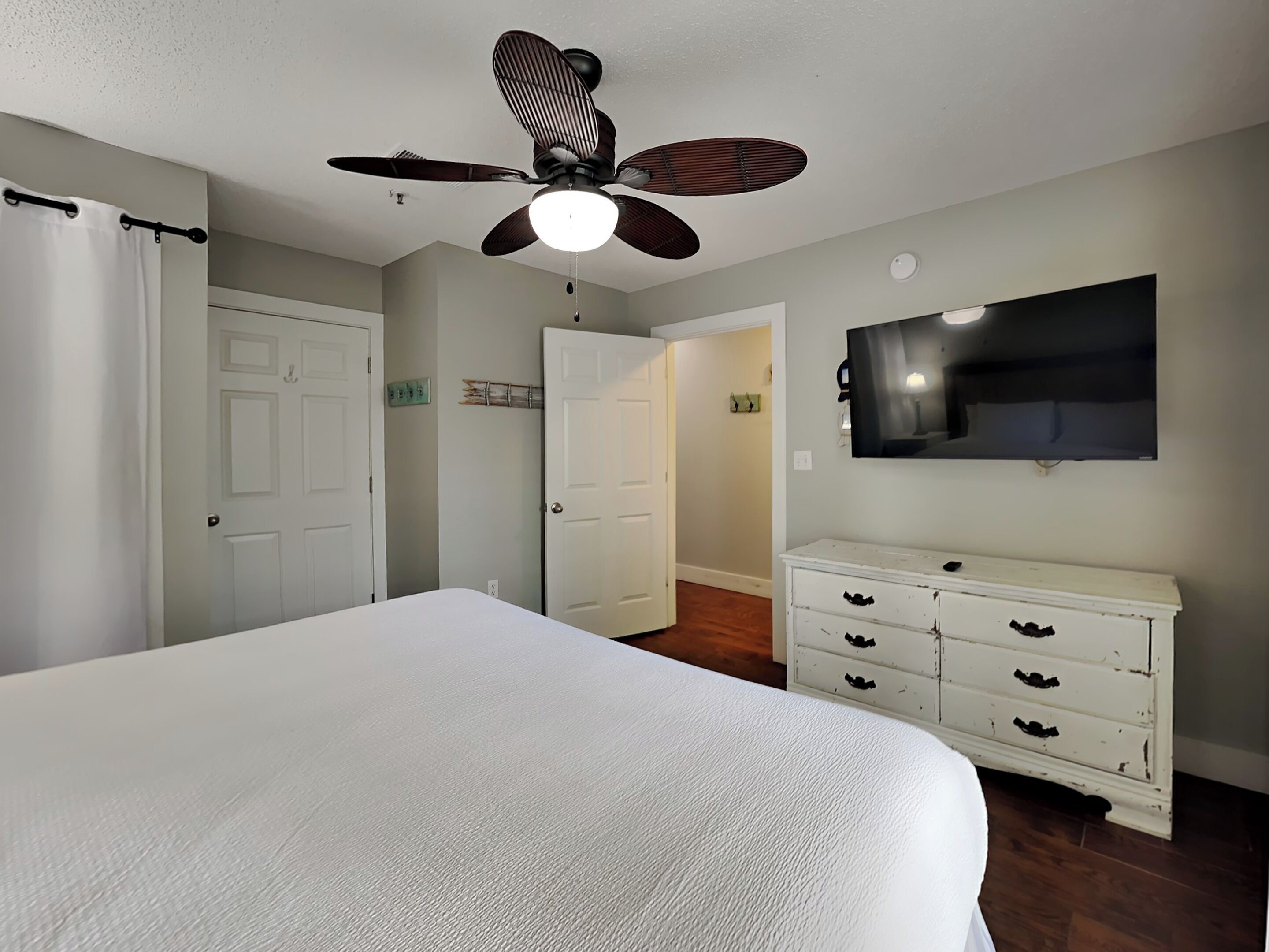 A bedroom with a bed, a ceiling fan, a dresser, and a TV mounted on the wall.