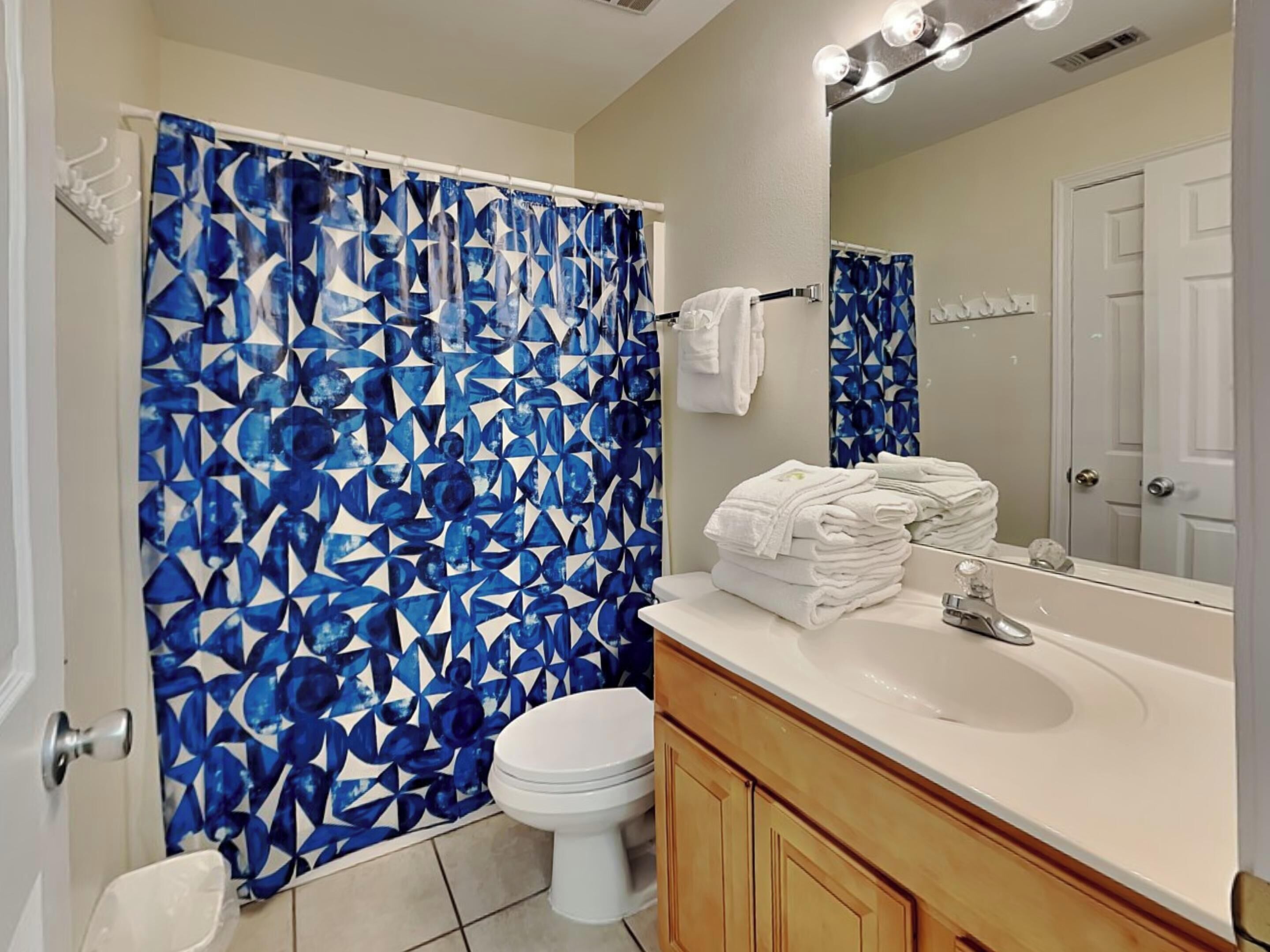A bathroom with a geometric blue shower curtain, a white toilet, a sink with a mirror, and neatly stacked towels.