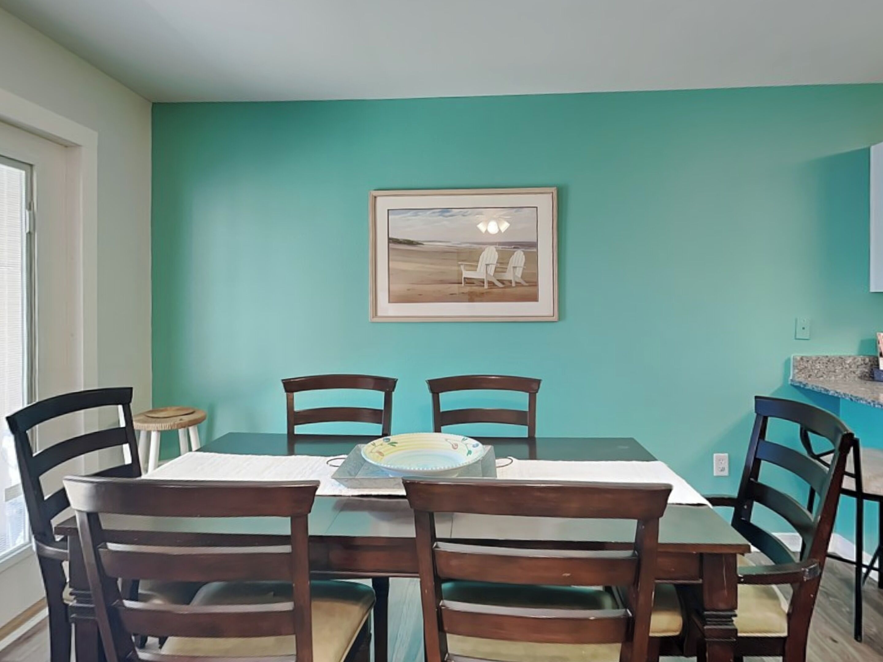 A dining area with a table set for four, two chairs on each side, a framed picture on the wall, and a small stool.