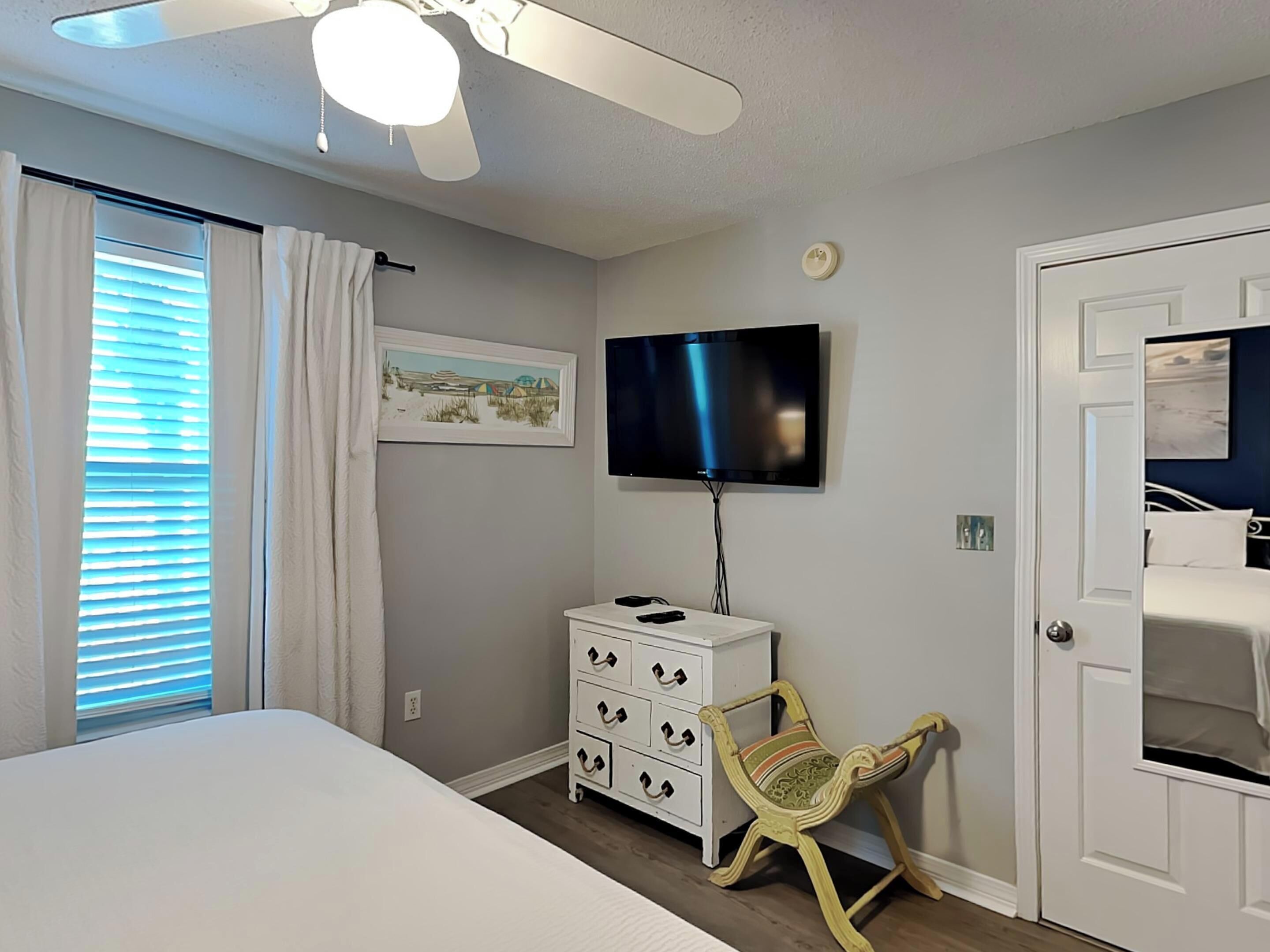 Quarto com cama, televisão na parede, cômoda, cadeira e porta.