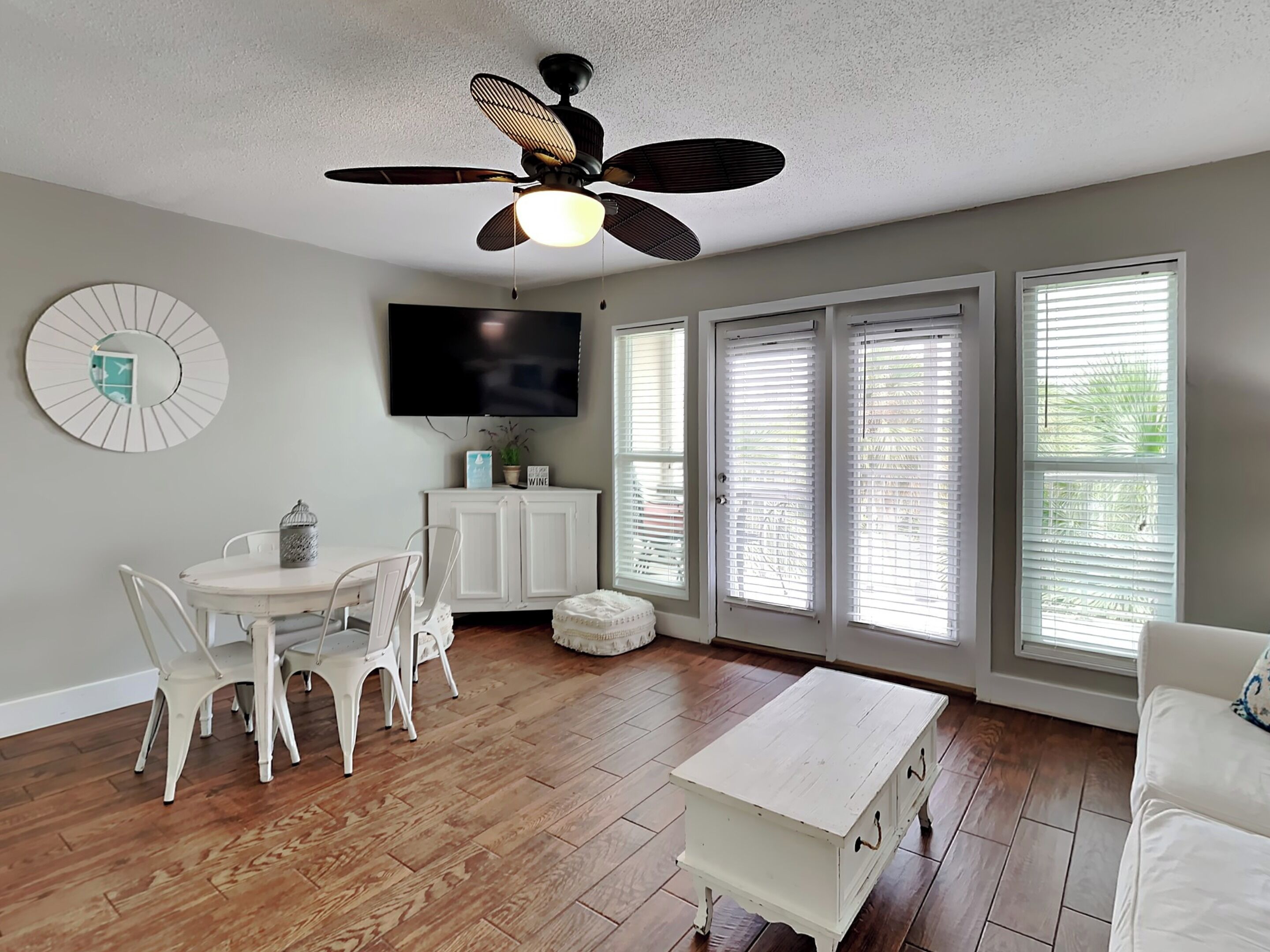 A spacious living room with a ceiling fan, a flat-screen TV, a dining area with white chairs, and large windows with blinds.