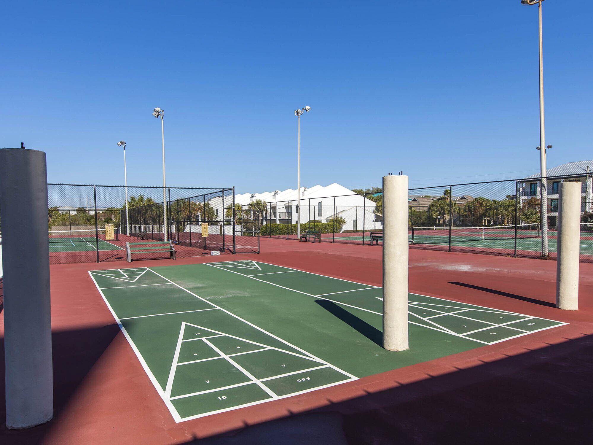 Un court de tennis extérieur doté d’une surface de jeu verte, entouré d’une clôture en treillis métallique et situé à côté d’un bâtiment à l’extérieur blanc.