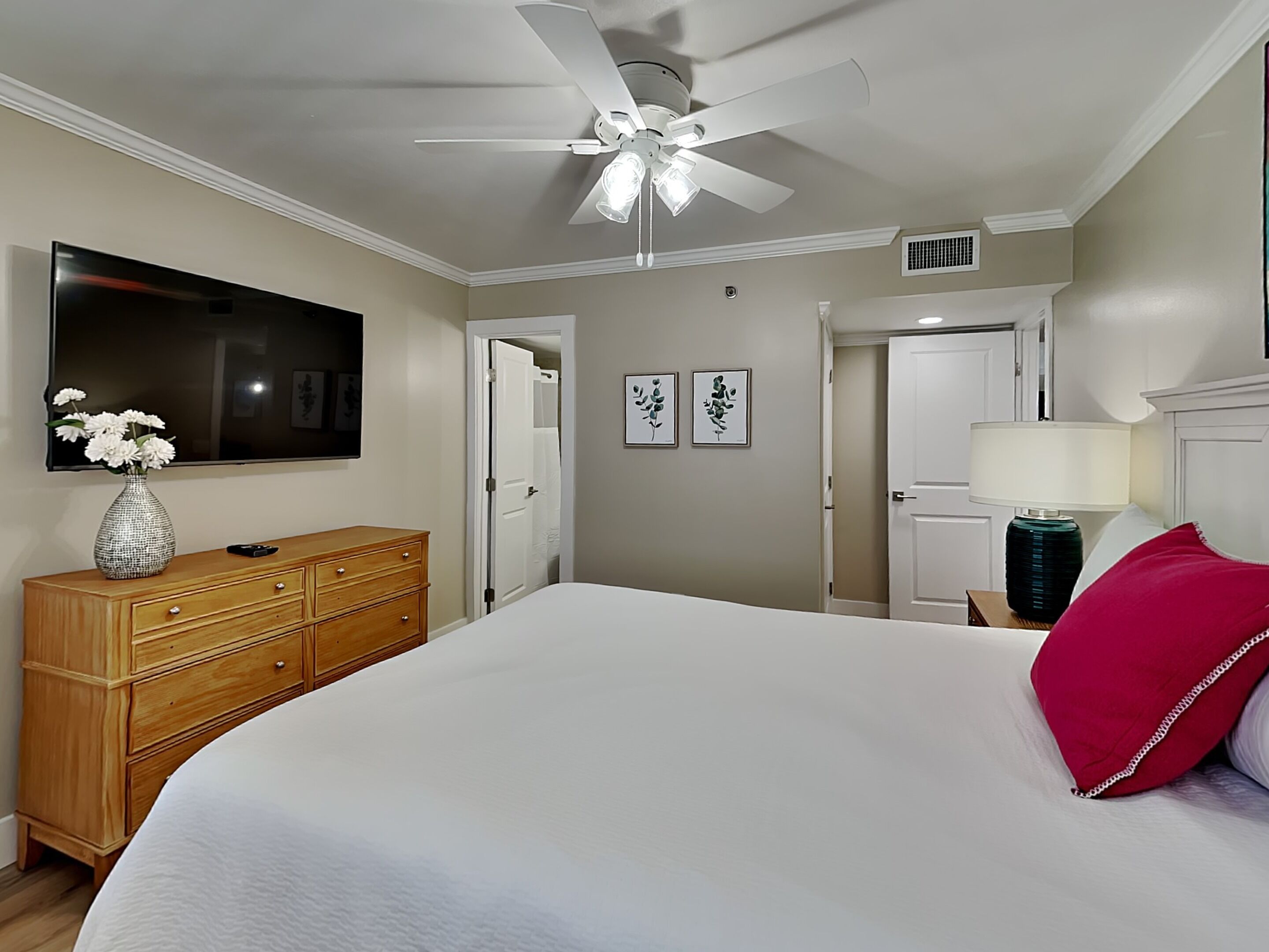 A bedroom with a bed, a wooden dresser, a TV, a ceiling fan, and two doors.