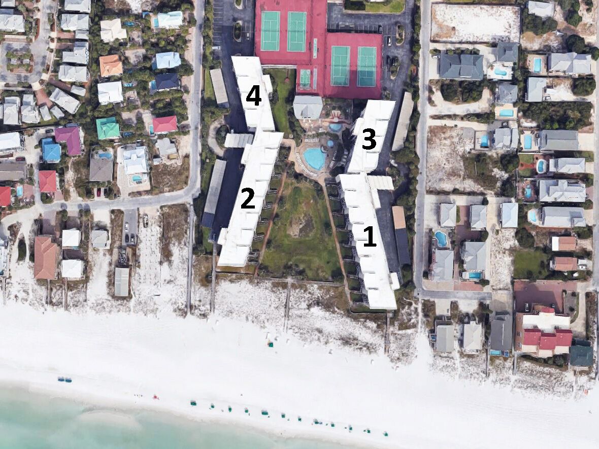 Vista aérea de uma área residencial costeira com edifícios numerados, uma praia e um prédio grande vermelho com janelas verdes.