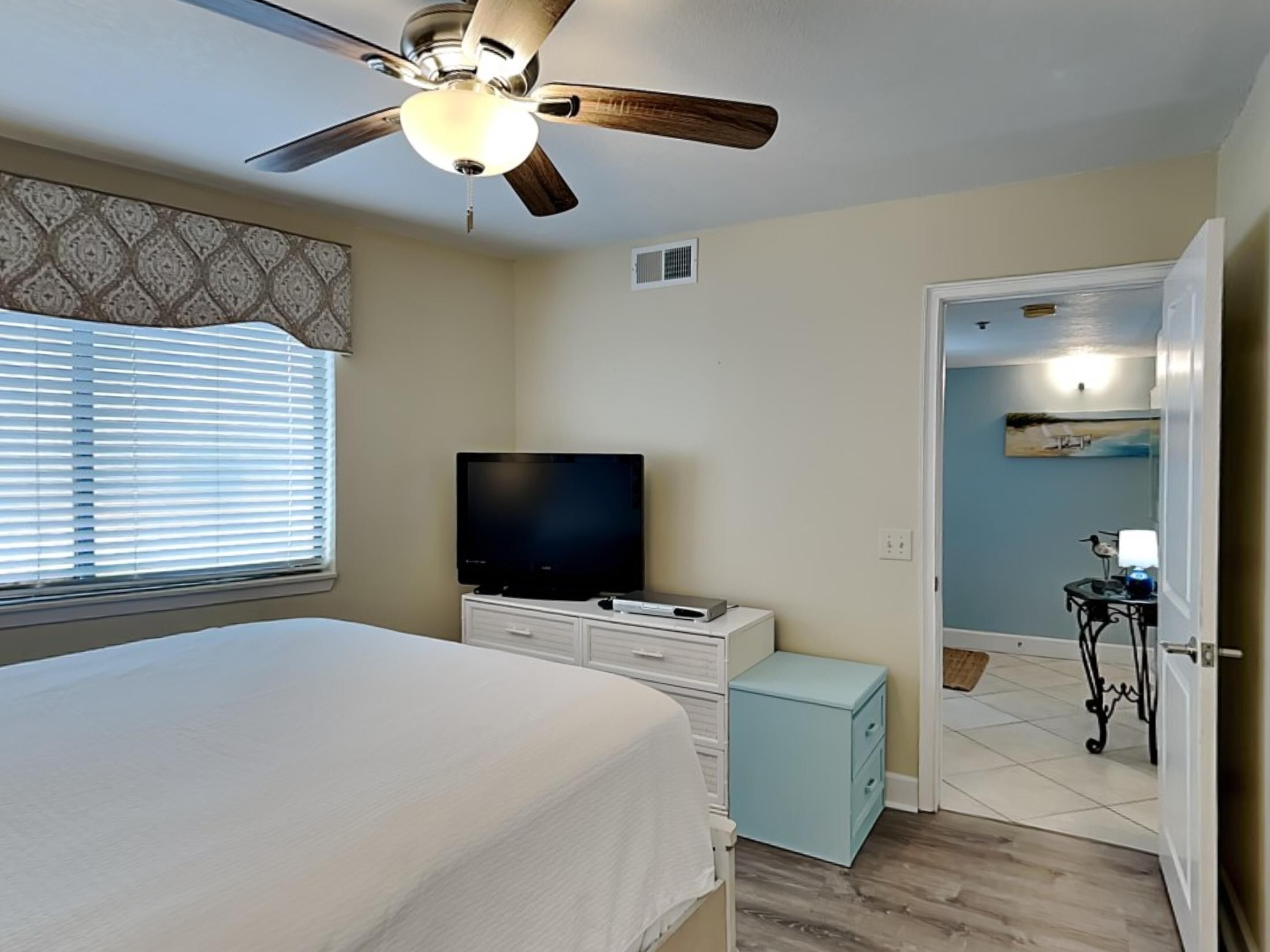 A bedroom with a bed, a ceiling fan, a television, a small cabinet, and a door leading to another room.