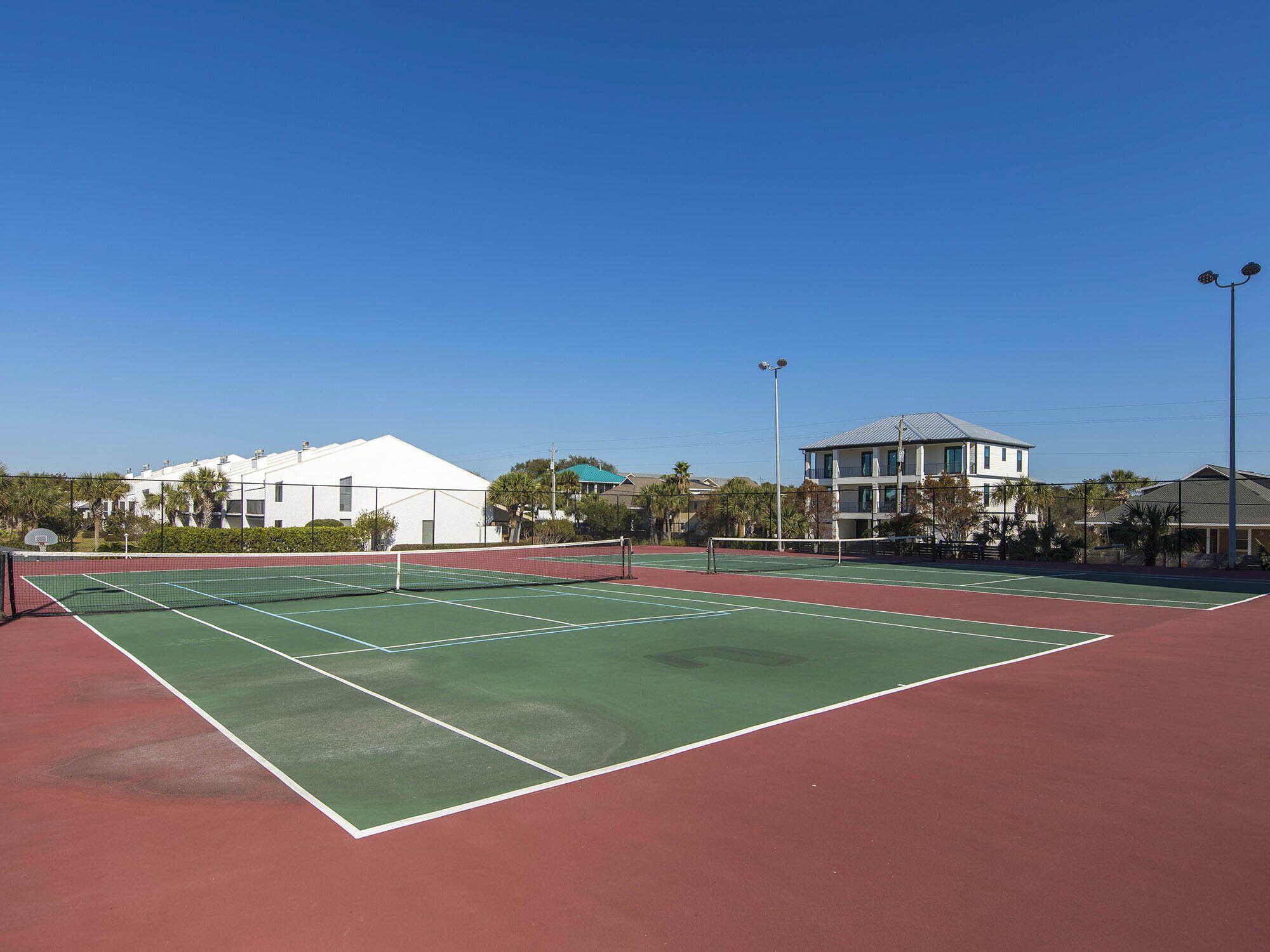 A tennis court with a net, surrounded by a fence, and buildings in the background.