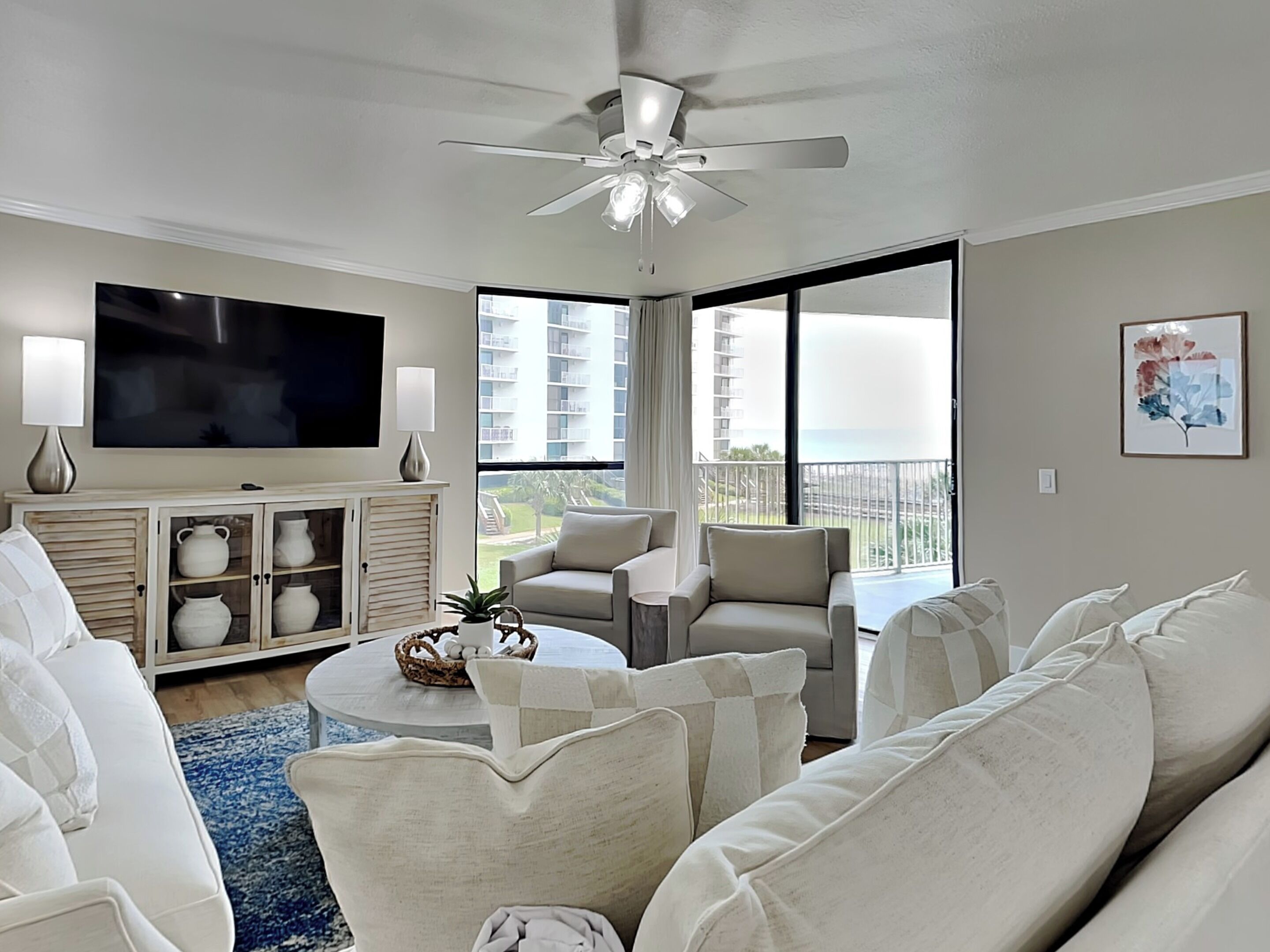 A spacious living room with a ceiling fan, a flat-screen TV, a large window with a view of buildings, and a balcony.