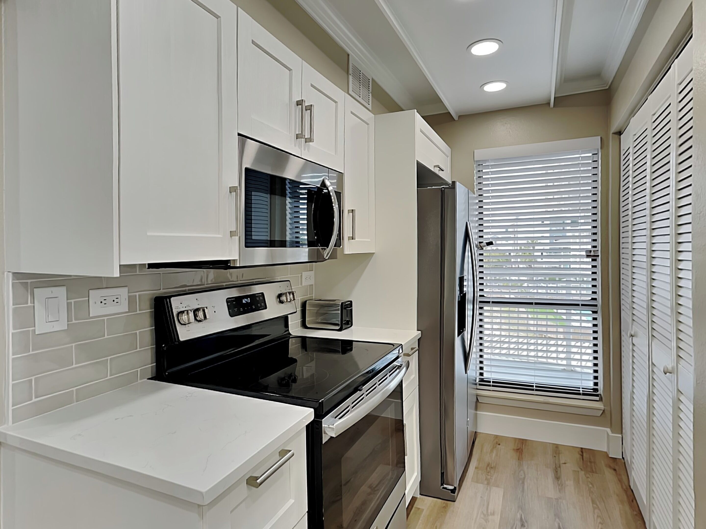 A modern kitchen with white cabinetry, stainless steel appliances, and a window with blinds.