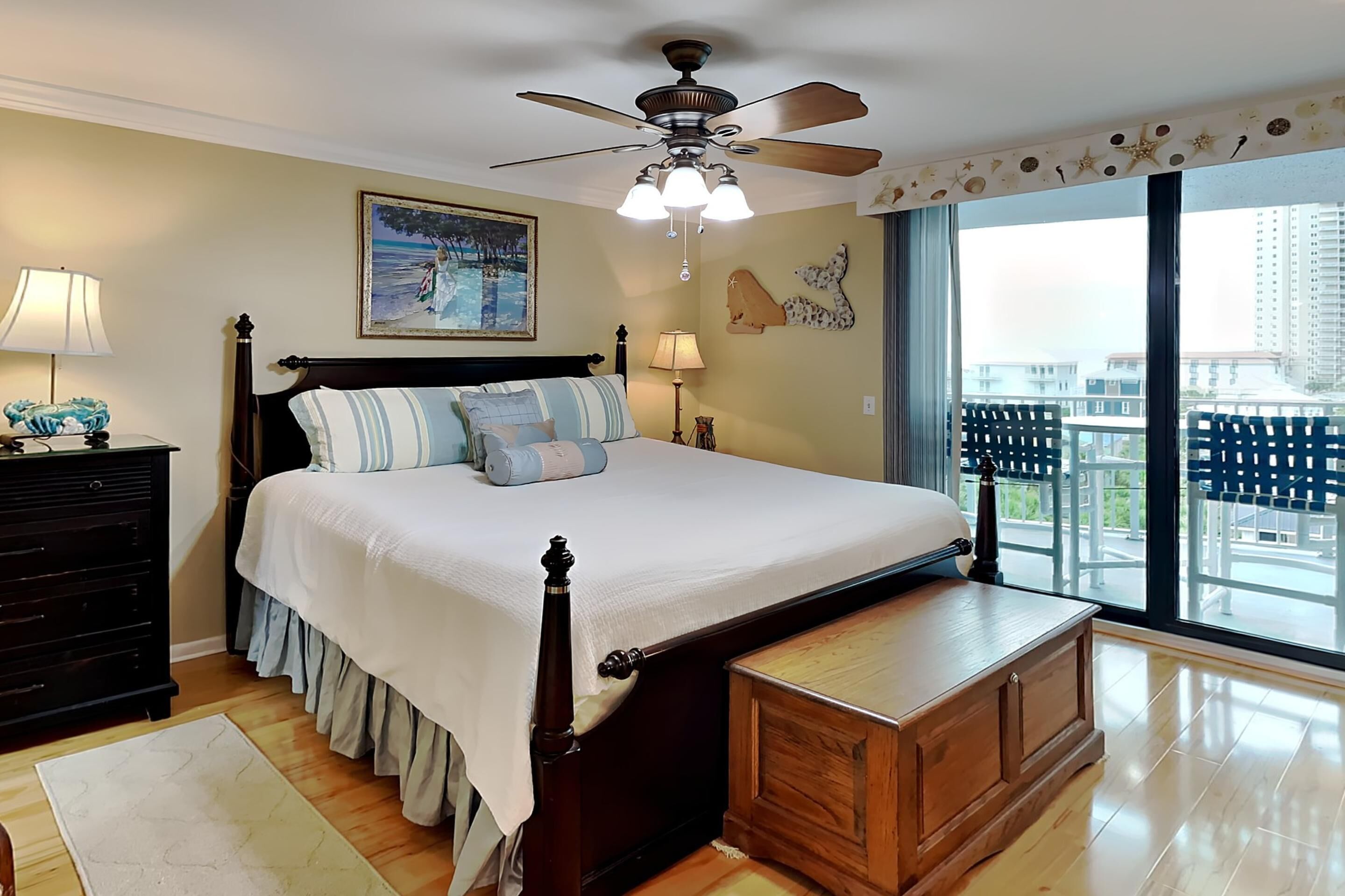 A bedroom with a large bed, a ceiling fan, a nightstand, a dresser, and a view of the city through large windows.