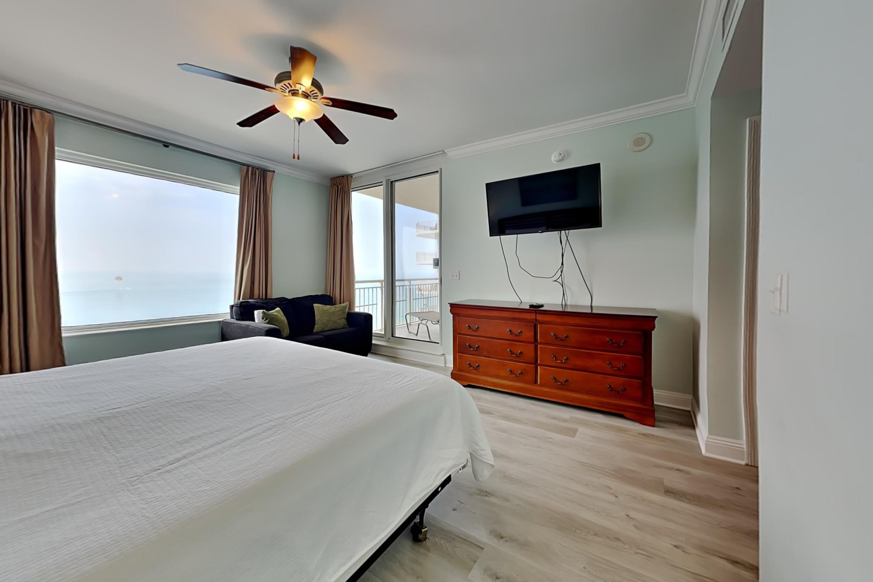 Une chambre avec un grand lit, un ventilateur de plafond, un téléviseur fixé au mur, une commode en bois et une vue par la fenêtre.