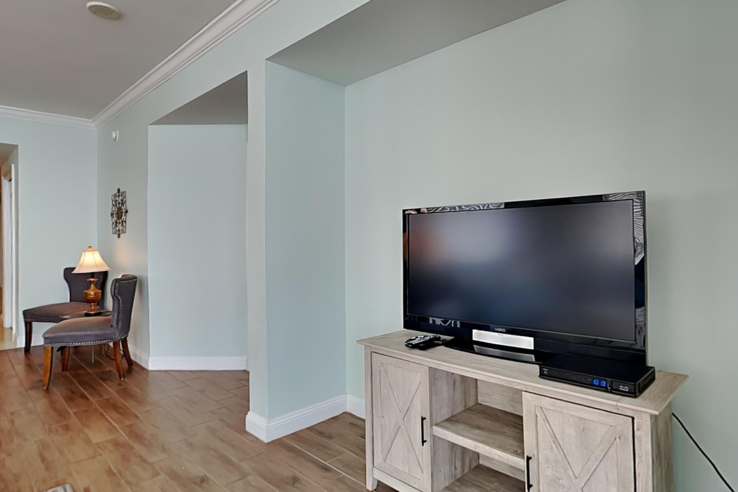 Un salon avec un téléviseur à écran plat posé sur un meuble en bois, un coin repas avec une table et des chaises, et un couloir avec une porte.