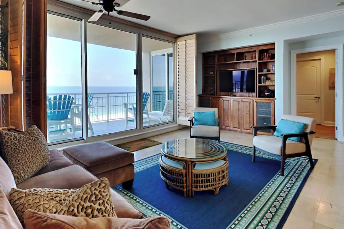 Un vaste espace de vie avec une grande porte-fenêtre coulissante donnant sur un balcon avec vue sur l’océan. On y trouve un coin salon confortable avec un canapé et des fauteuils, une petite table basse ronde et une bibliothèque.