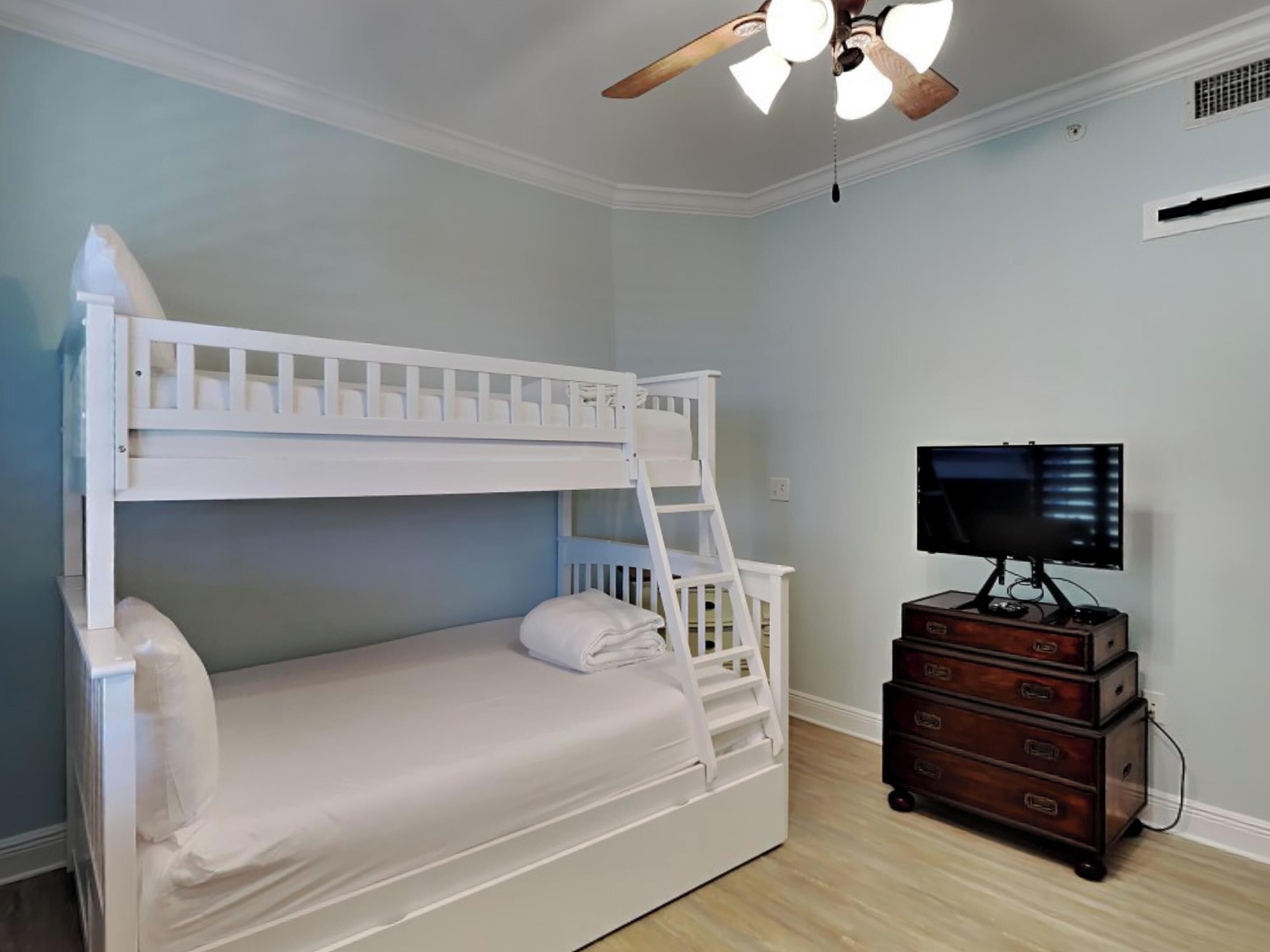 Quarto com beliche, ventoinha no teto, televisão em cômoda de madeira e paredes azul-claras.