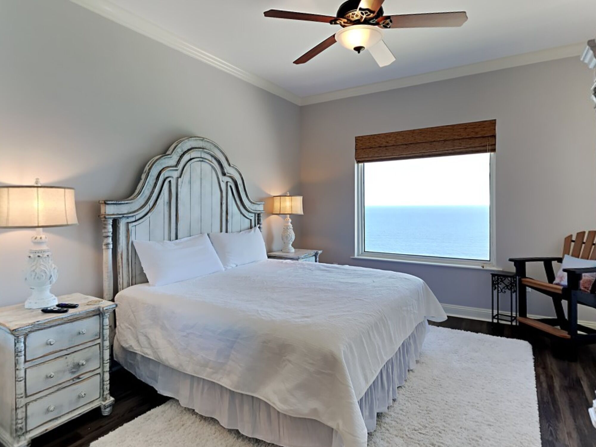 A bedroom with a large bed, two bedside tables, a ceiling fan, a window with a view of the ocean, and a wooden chair.