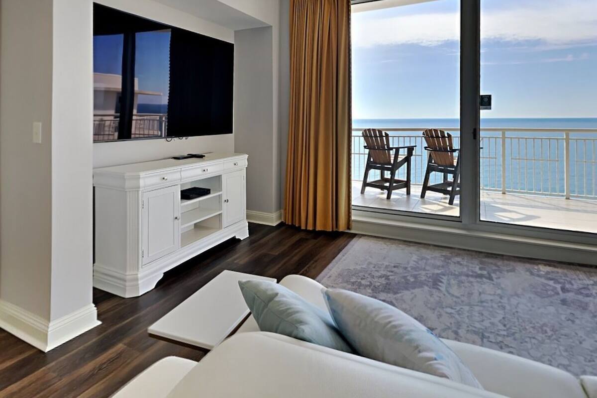 Un salon moderne doté d’un téléviseur à écran plat, d’un meuble de rangement blanc, d’un canapé avec des coussins et d’un balcon offrant une vue sur la mer.