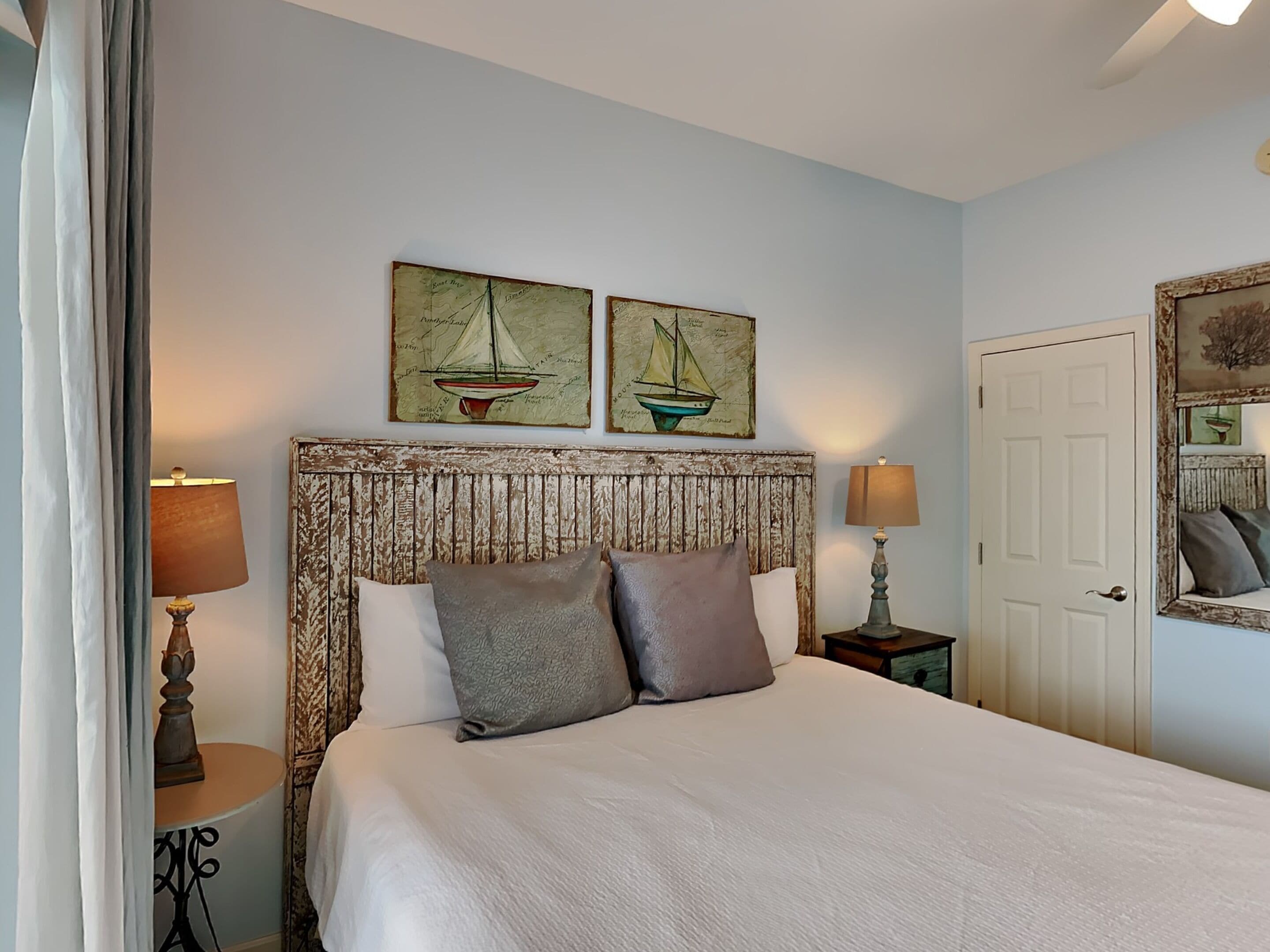 A bedroom with a large bed, two wall-mounted paintings of sailboats, a rustic headboard, bedside tables with lamps, and a door with a mirror.
