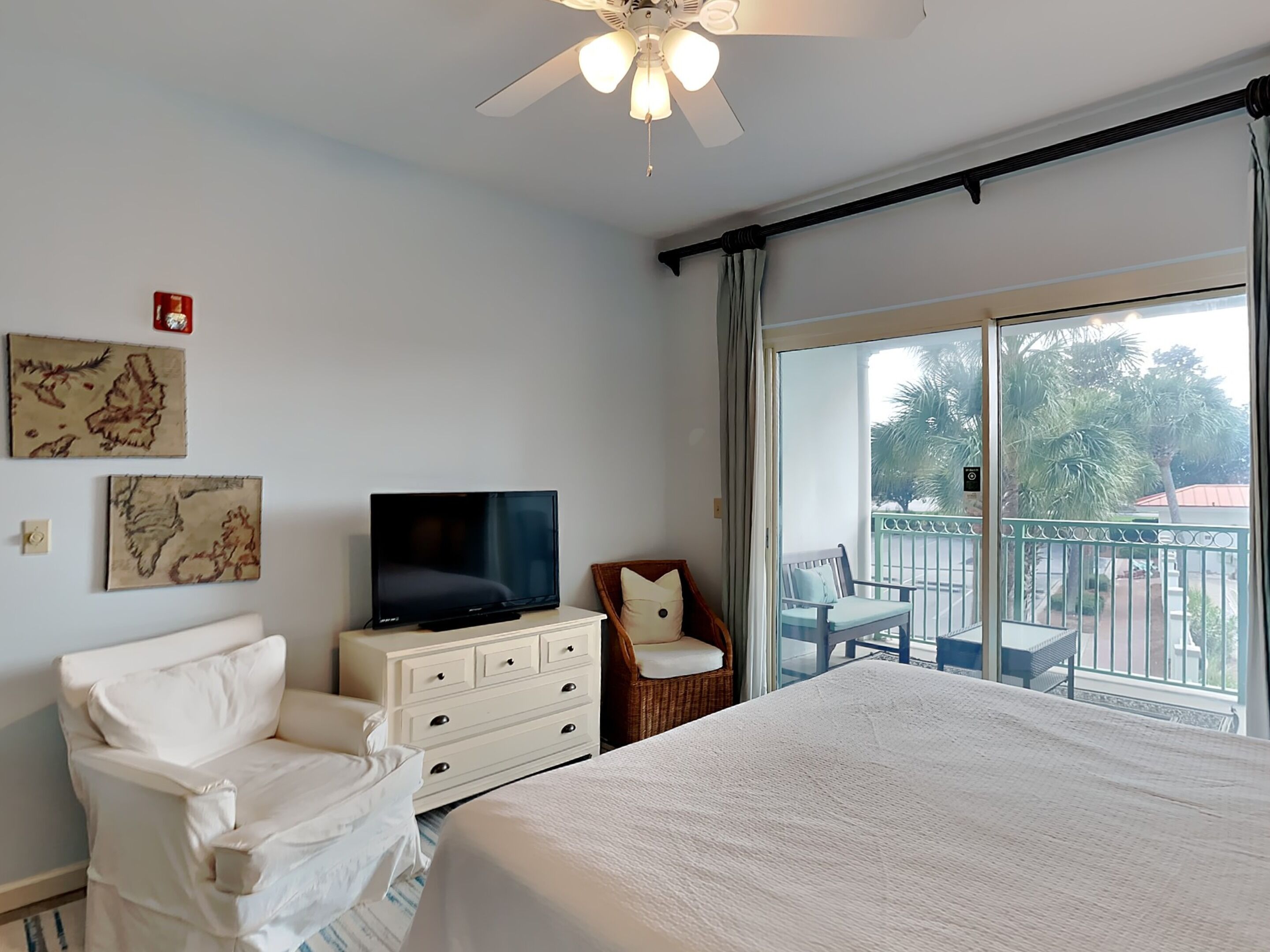 Habitación de hotel con una cama, televisión, tocador blanco, sillón blanco y un balcón con vistas a palmeras.
