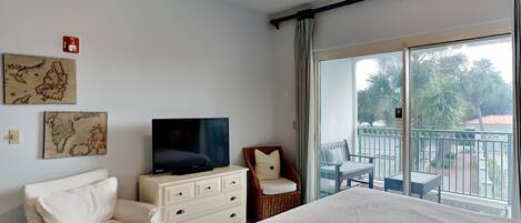 Habitación de hotel con una cama, televisión, tocador blanco, sillón blanco y un balcón con vistas a palmeras.