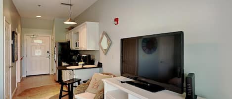 A living room with a television, white furniture, and a kitchen in the background.