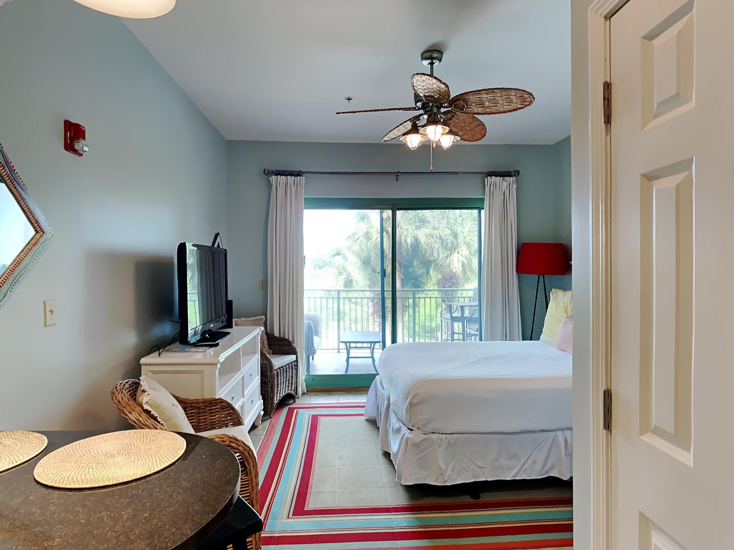 Une chambre à coucher comprenant un lit, une télévision, un ventilateur de plafond, un balcon avec vue sur les palmiers et un tapis à rayures.
